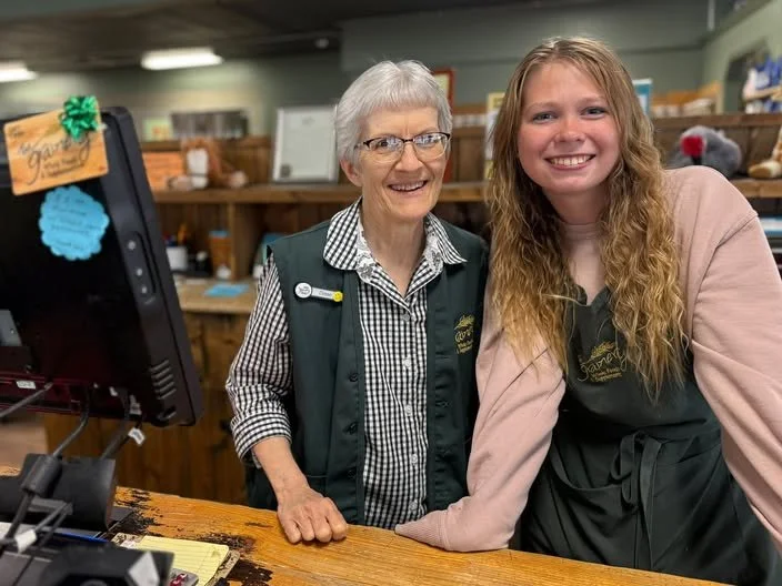 An older woman with glasses and gray hair standing behind a checkout counter with a young woman with long curly hair. Both are smiling. The background has wooden shelves and a computer on the counter.