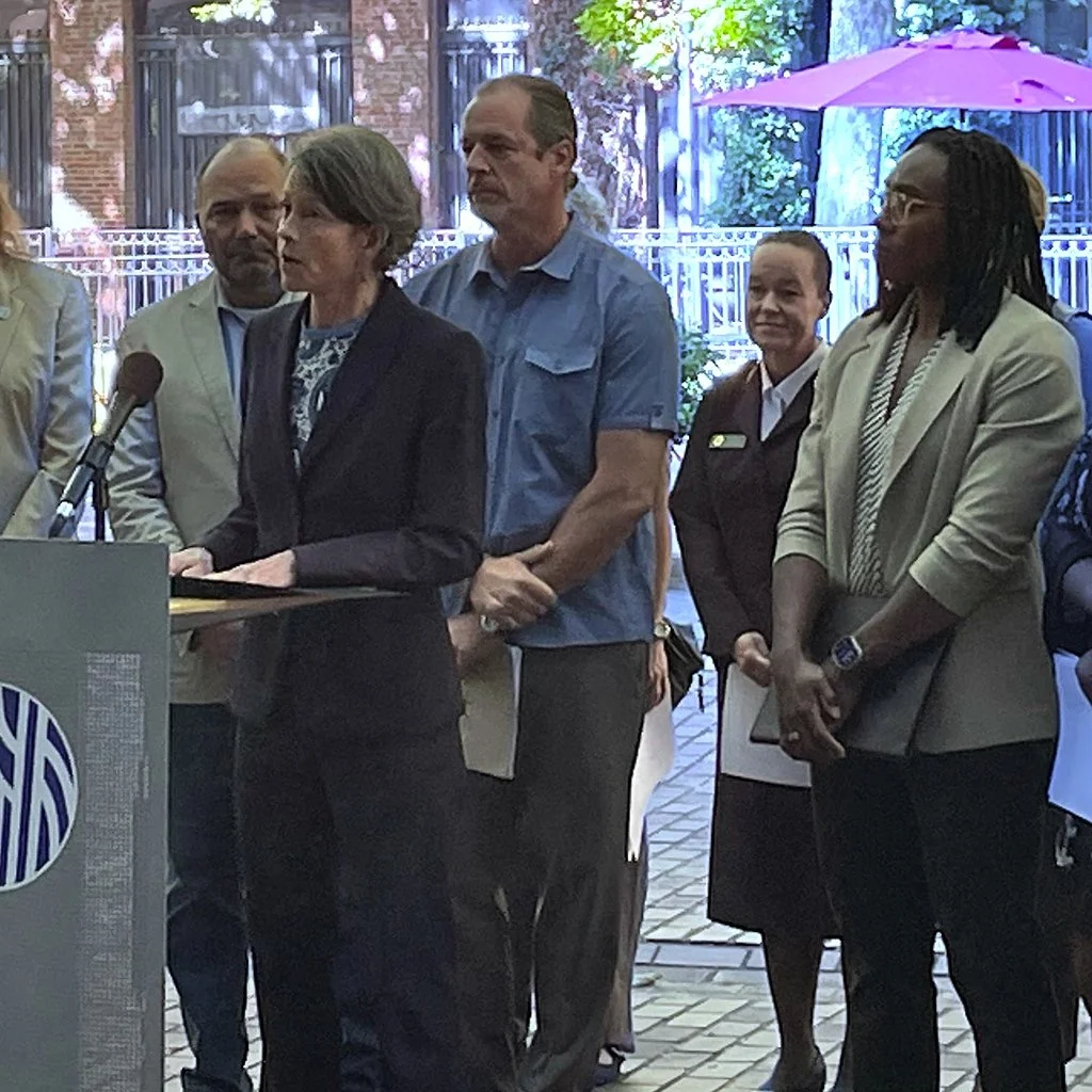 Sara Nelson speaks in a black blazer is speaking at a podium with five other people standing behind her, outdoors with a wire fence, trees, and an umbrella visible in the background.