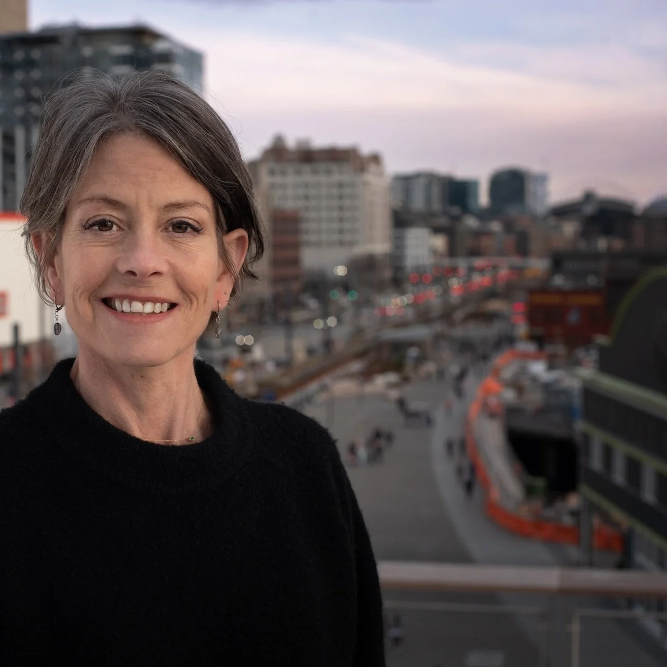 Sara Nelson, with short gray hair wearing a black top, smiling against a cityscape backdrop at dusk with the Seattle waterfront in the background.