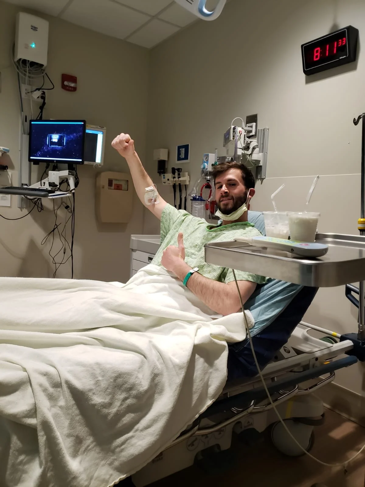 A man in a hospital bed giving a thumbs-up, with a fist raised, wearing a hospital gown and a face mask pulled down. There are medical equipment and monitors around him, including a digital clock showing 8:11 PM.