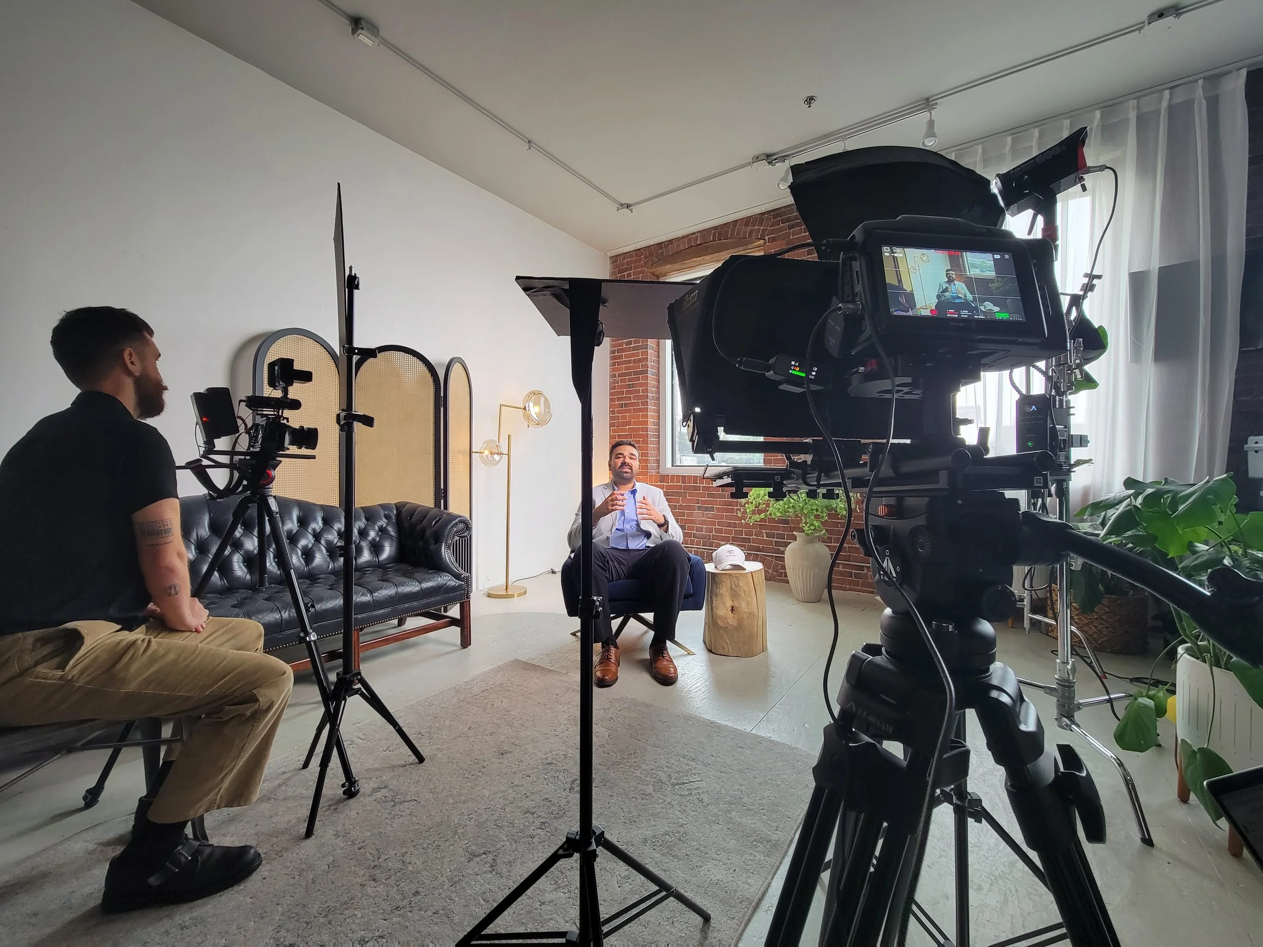 A man in a business suit being interviewed on camera in a modern, well-lit room with brick walls, plants, and minimalist decor.