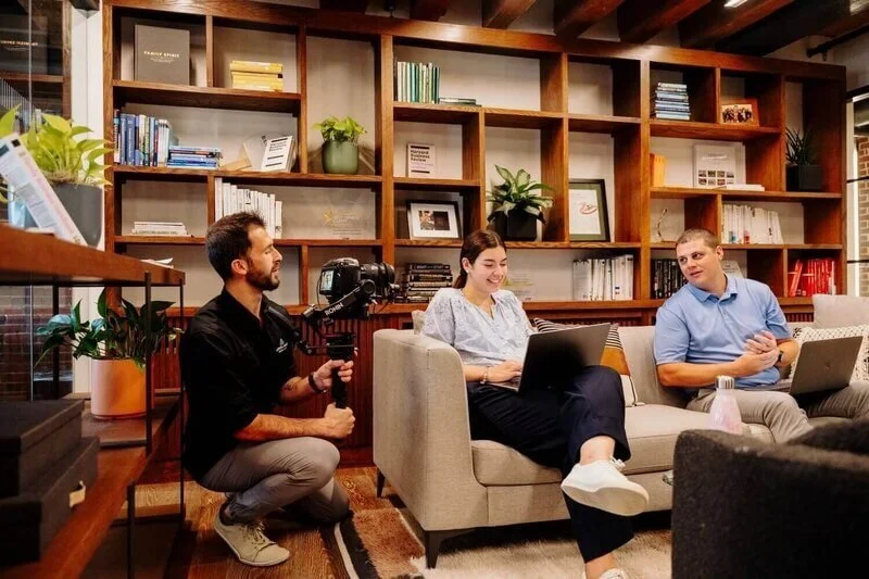 A man filming a woman and a man sitting on a couch with laptops in a cozy room with wooden shelves, books, and plants.