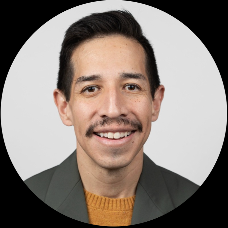 Portrait of a man with dark hair, mustache, and wearing a dark blazer and orange sweater, smiling against a plain light background.