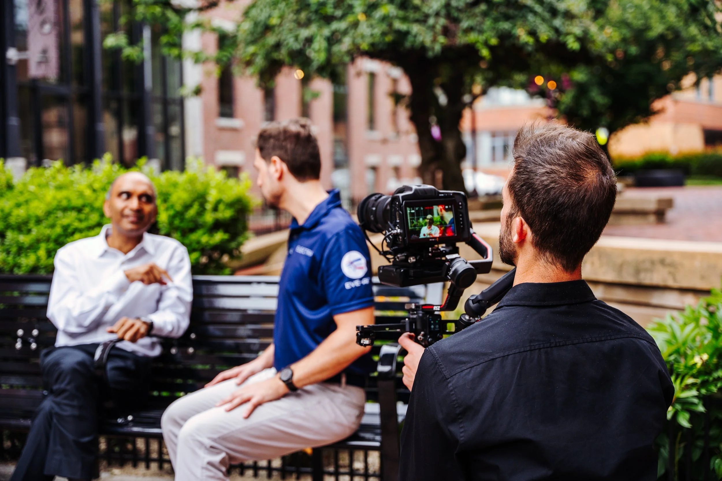 A man operating a camera filmed two men having a conversation on a park bench outdoors, with trees and buildings in the background.