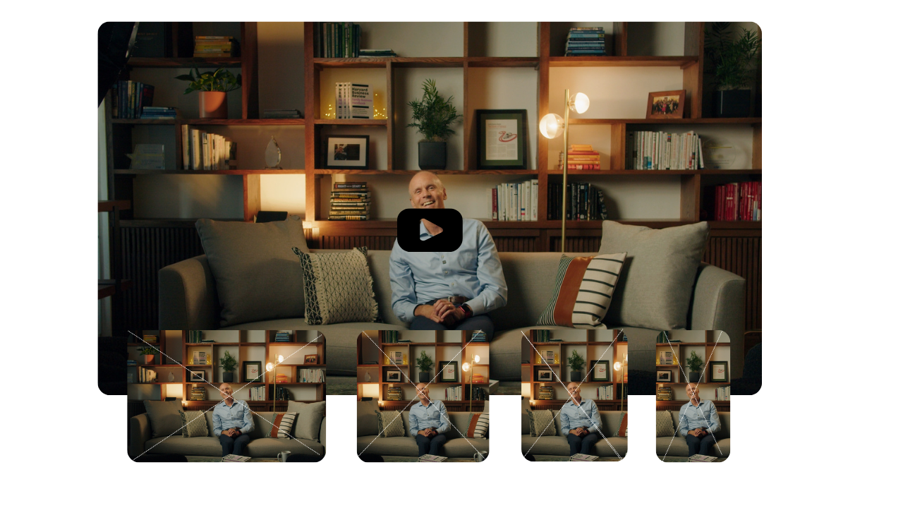 A man sitting on a gray couch in a living room, surrounded by pillows, with a wall of shelves filled with books and decorative items behind him. A standing lamp with multiple round bulbs is beside him.