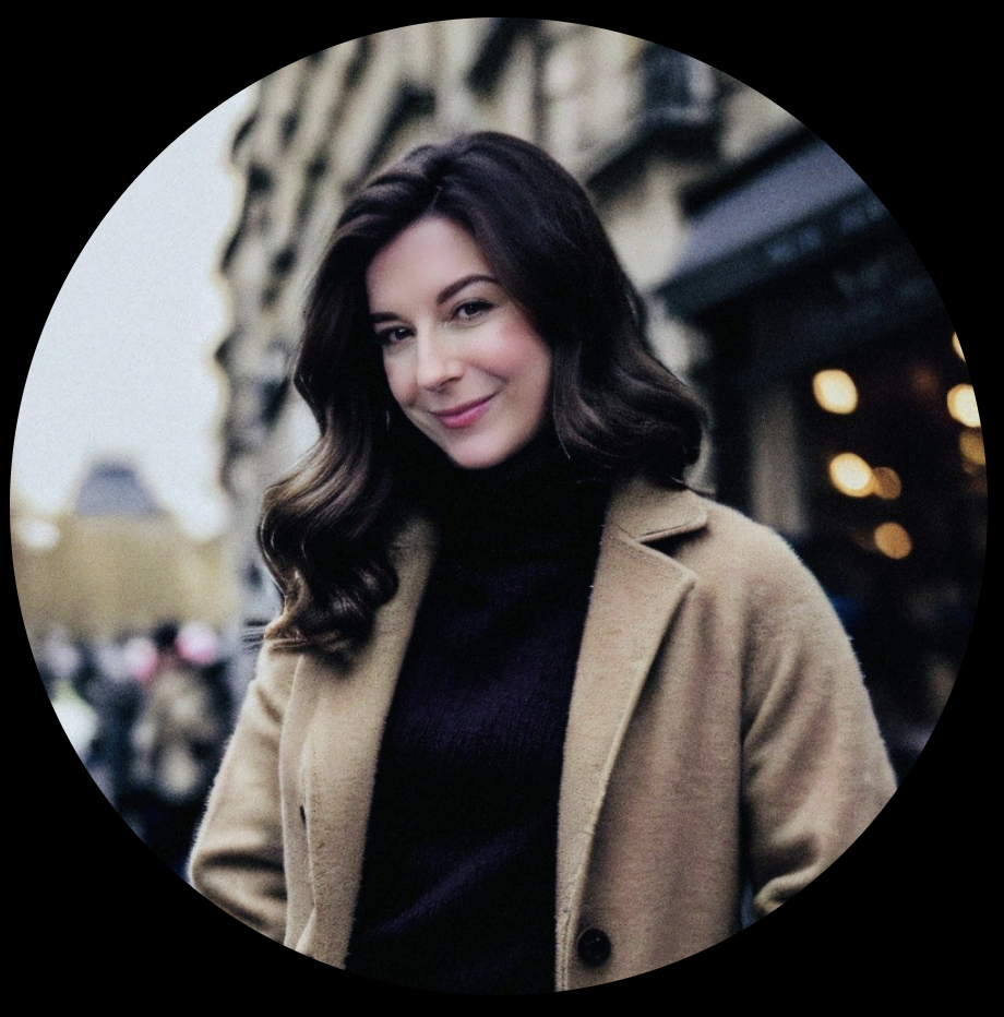 A woman with wavy dark hair smiling in an outdoor urban setting during evening, wearing a beige coat and black turtleneck.