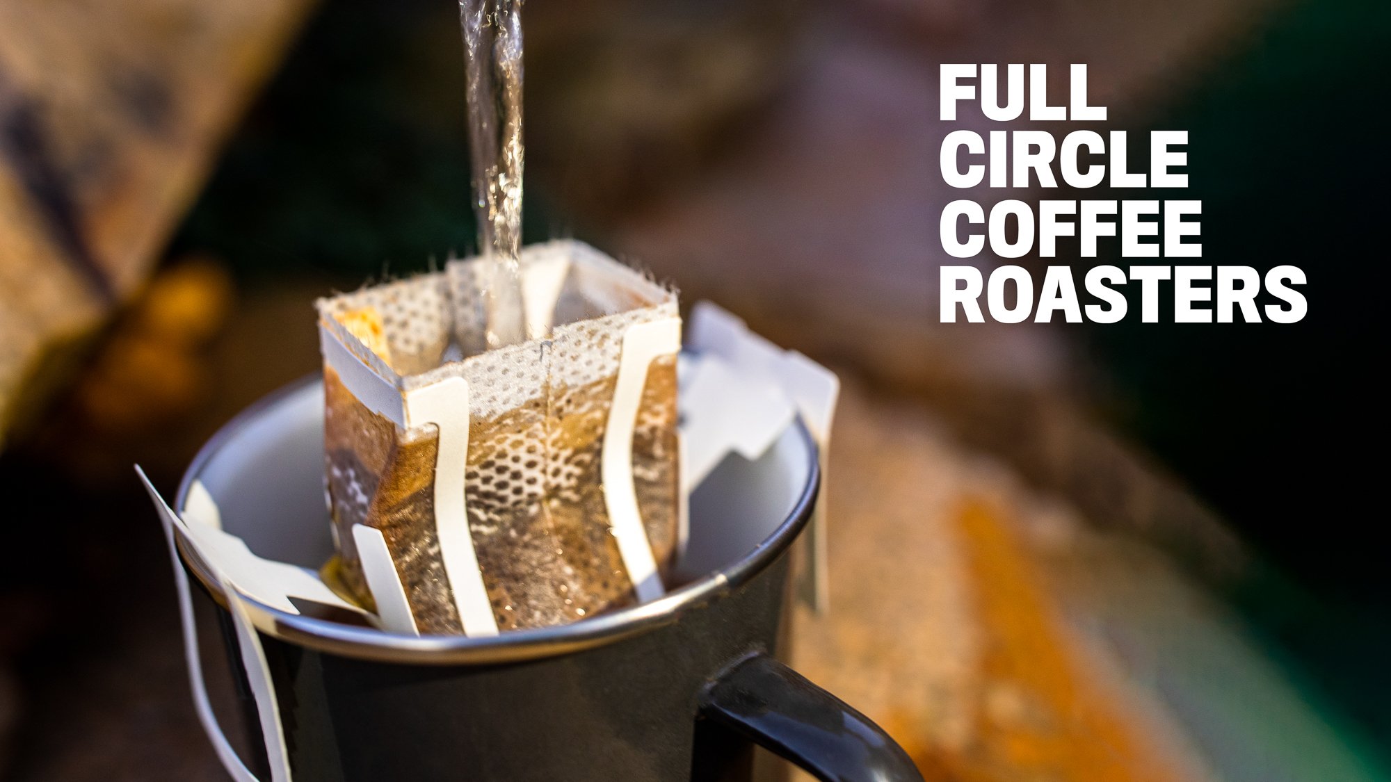 Pour-over coffee being brewed with a drip coffee bag over a black mug, with water being poured into the filter. The background has the text "Full Circle Coffee Roasters." by Justine Brentnall | Arty Crow Design