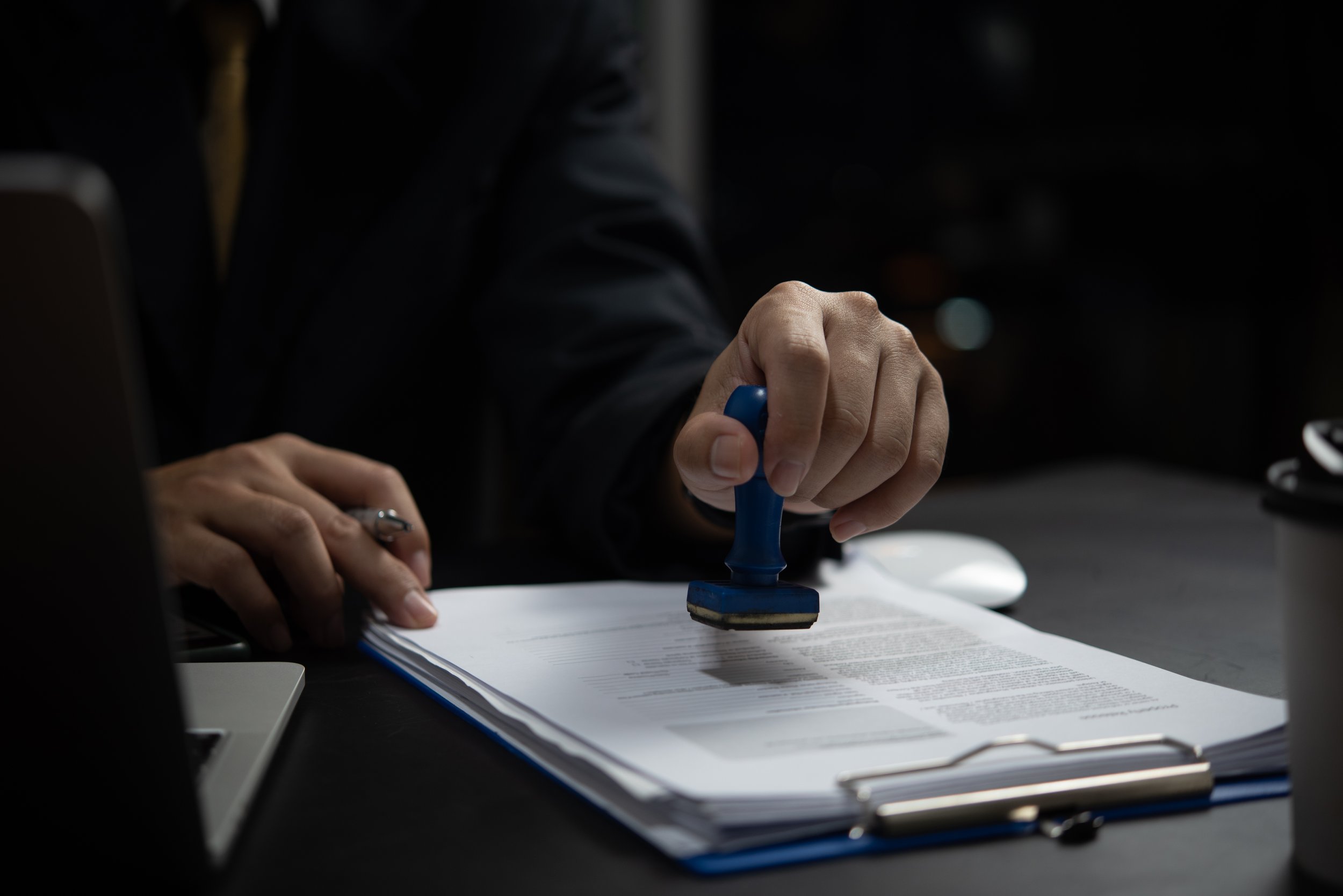 Person using a blue rubber stamp to mark or sign a document on a clipboard in a dark setting.