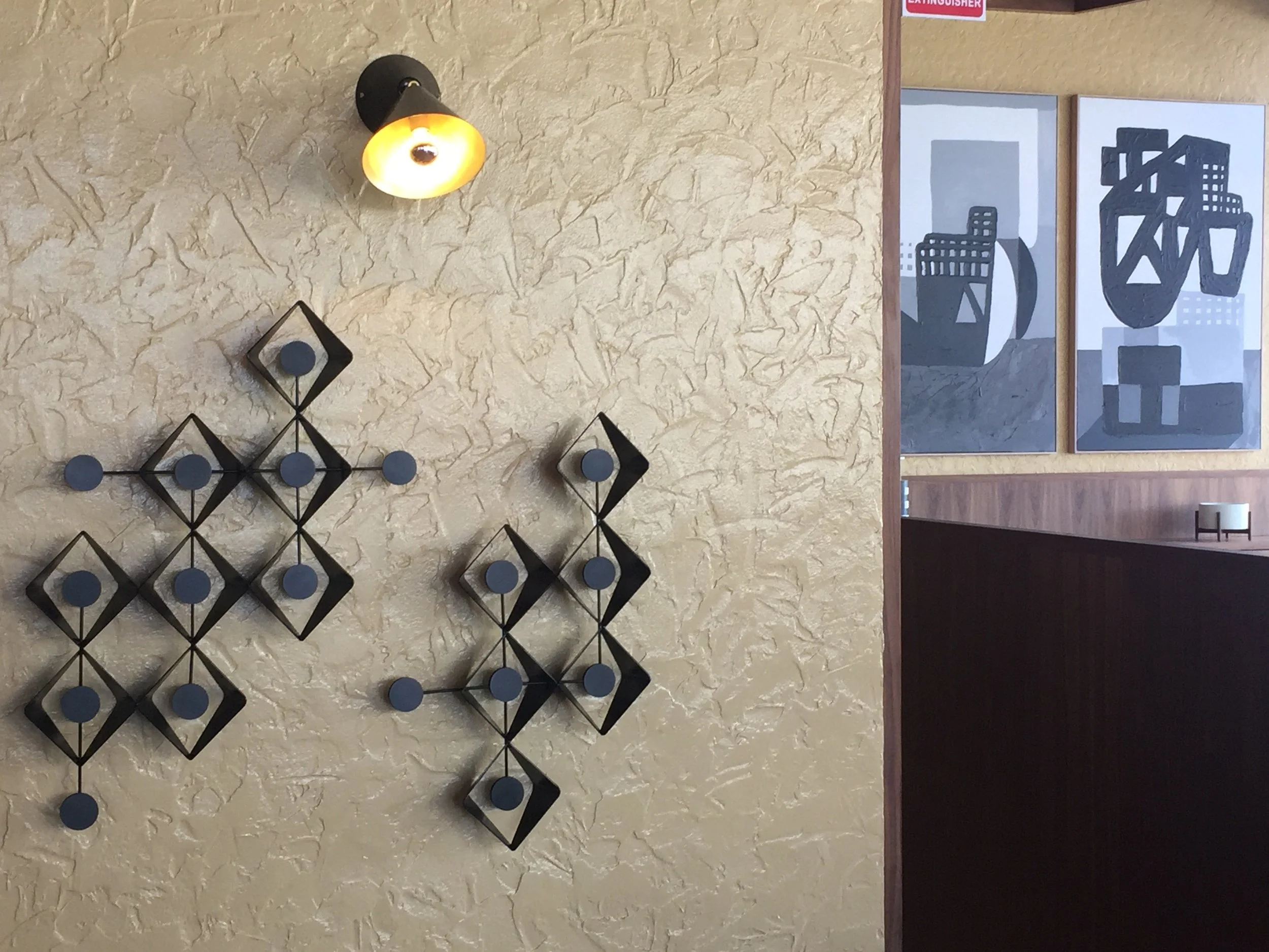 Decorative black metal wall art with circular accents on a textured beige wall, illuminated by a small wall sconce with a yellowish light. To the right, part of a wooden partition with two framed abstract cityscape artworks in shades of gray and black.