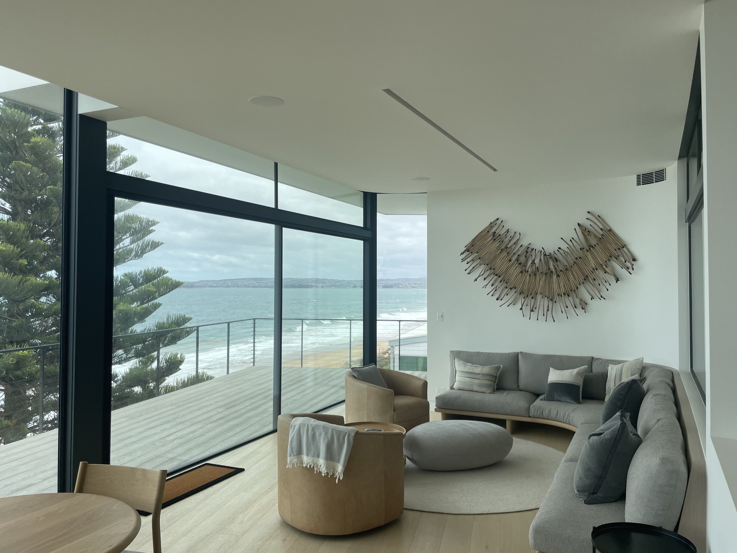 Modern living room with large glass windows overlooking the ocean, featuring a grey sectional sofa, a beige armchair, a round ottoman, and wall art made of arranged spoons.
