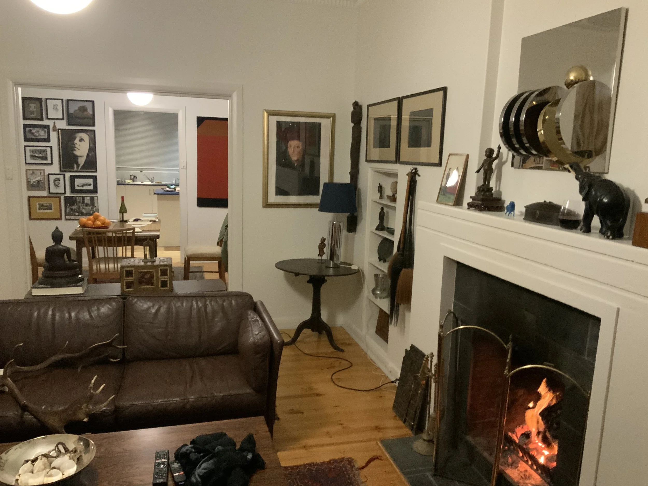 A cozy living room with a lit fireplace, a leather sofa, a side table with art pieces, and decorated walls. chairs, a bowl of oranges, and a bottle of wine is visible through a doorway. curation, art install, sculpture install, sculpture hanging.