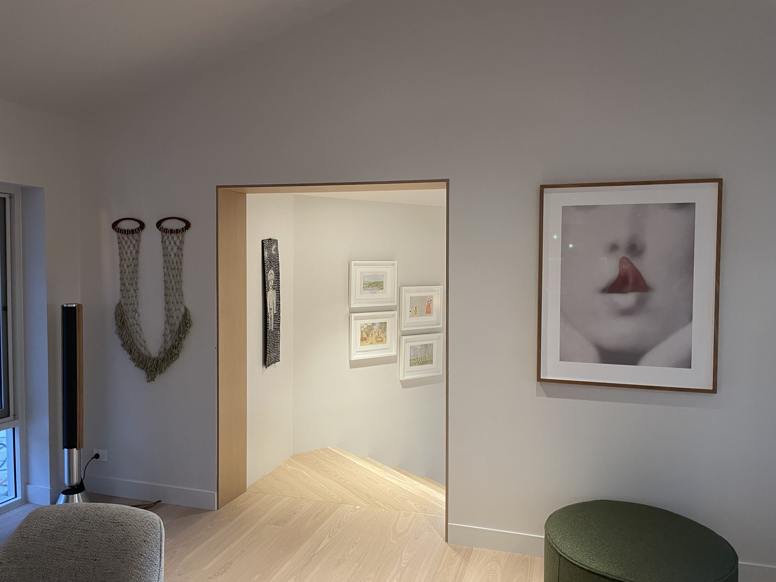 Interior view of a modern home with artwork on white walls, including framed pictures and a large black-and-white portrait of a face with lips and tongue visible, a woven wall hanging, and a sculptural wooden hand, with a green ottoman and a beige armchair in the foreground.