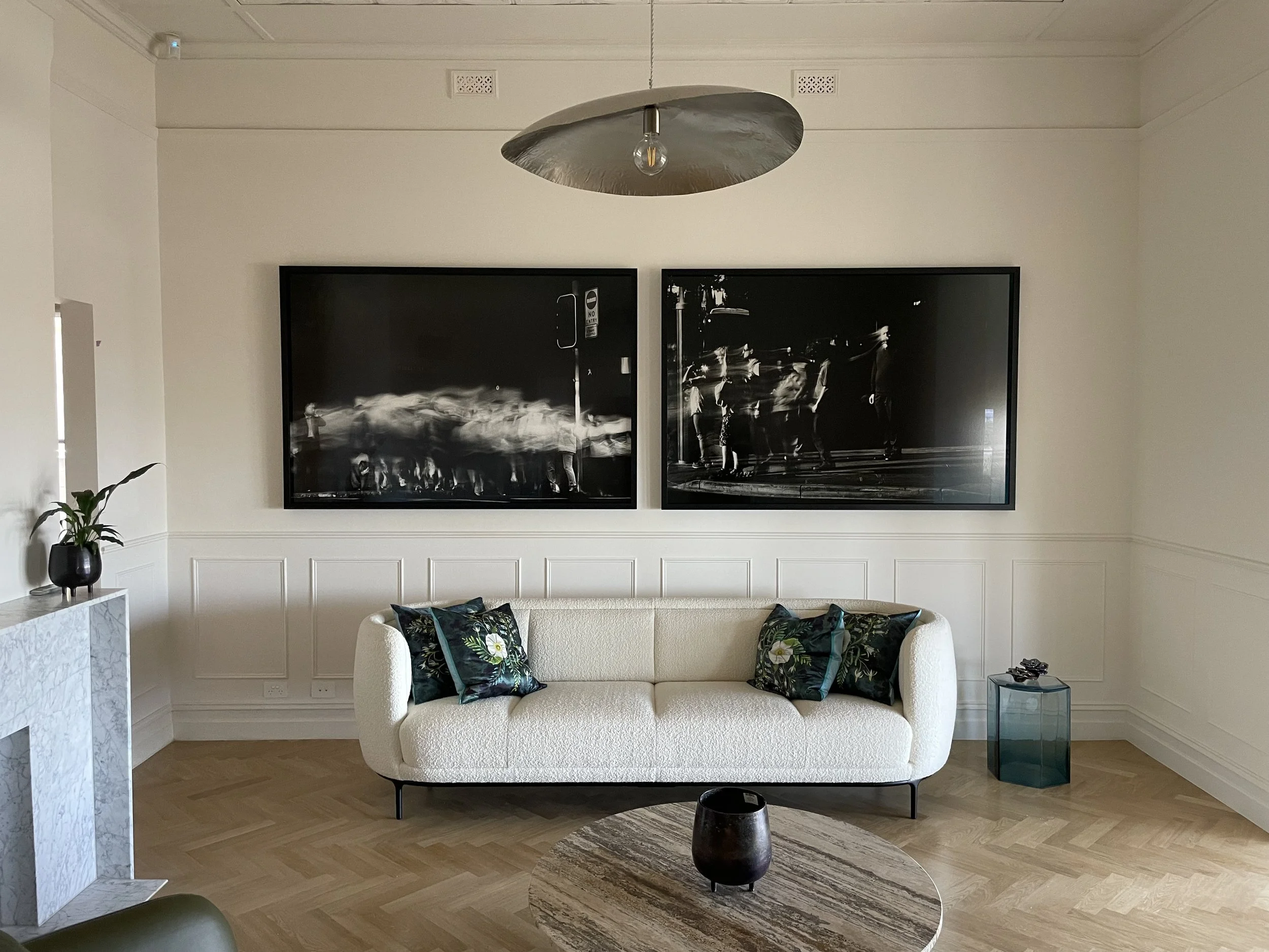 a round wooden coffee table, a small black vase in front, a dark green side table with object, a potted plant on the left, two large black-and-white photographs on the wall, a metal pendant light fixture, and wood flooring. Art hanging, art install.