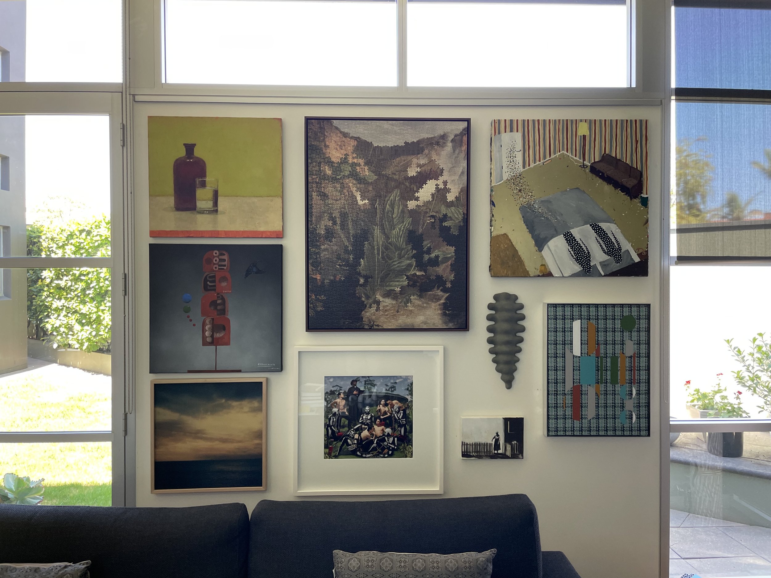 Living room wall with various framed artwork, including abstract paintings, landscape, and photographs, with sunlight coming through windows on either side. gallery style hang, art installation, picture hanging, sculpture install, sculpture hanging.