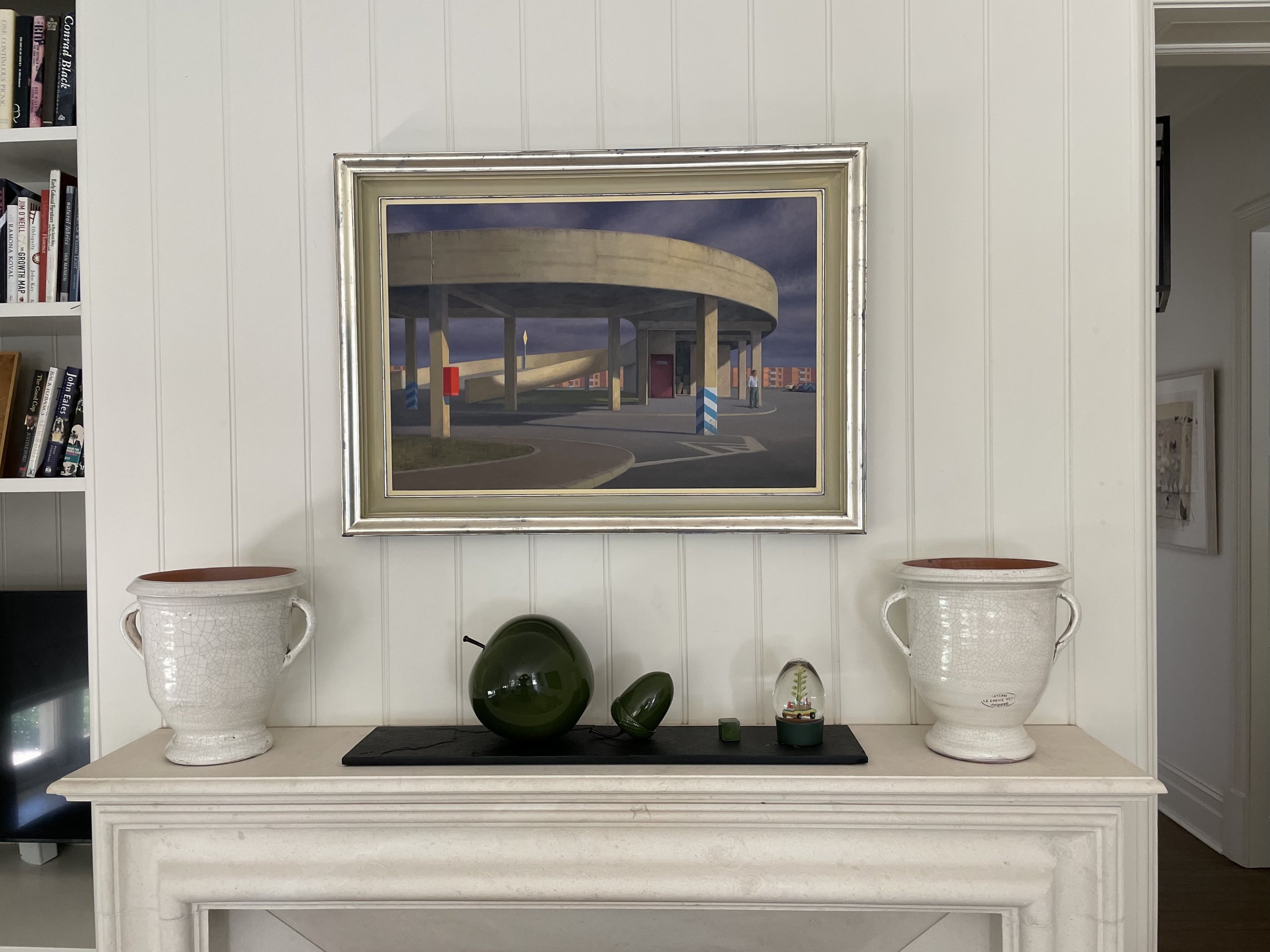 A framed painting of a concrete overpass with supporting columns, under a cloudy sky, hangs above a white fireplace mantel decorated with two large white ceramic vases. install of art work, art work hanging, art installation, gallery hang.