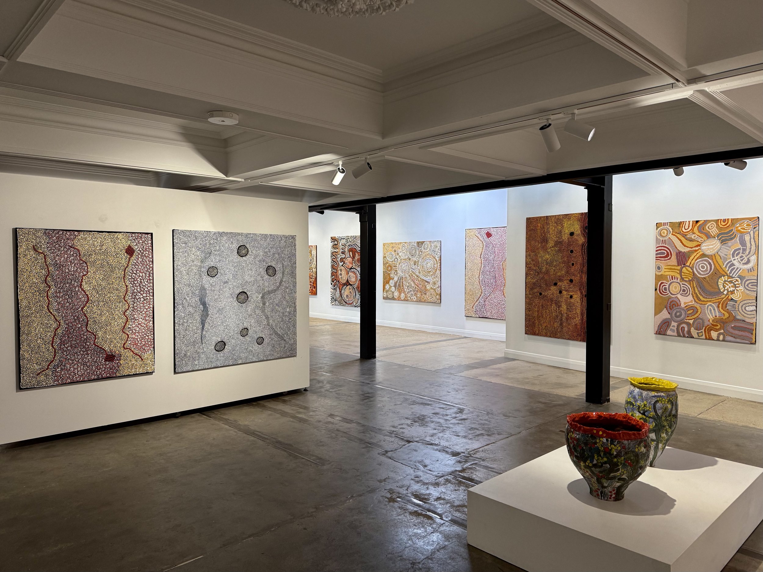 Interior view of an art gallery with colorful abstract paintings on the walls and decorative pots on a white pedestal in the foreground.