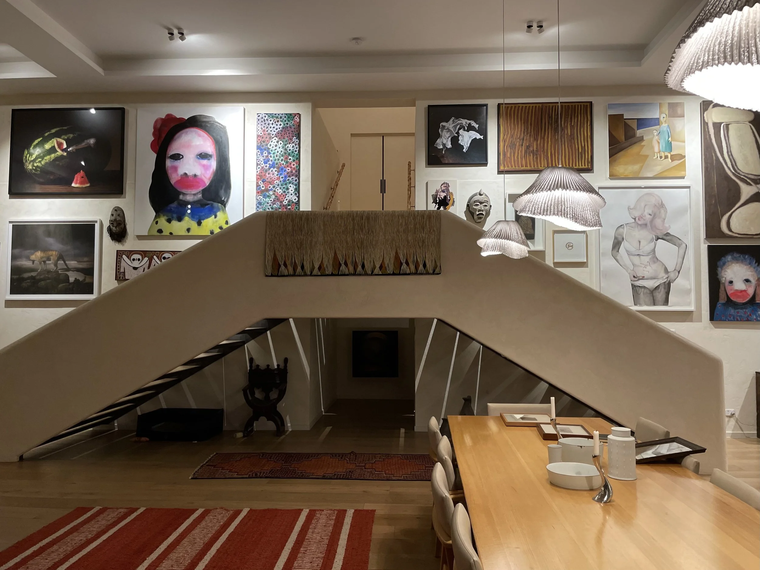 Interior of an art gallery with various paintings and masks on the wall, a staircase in the center, dining table and chairs, and hanging pendant lights.