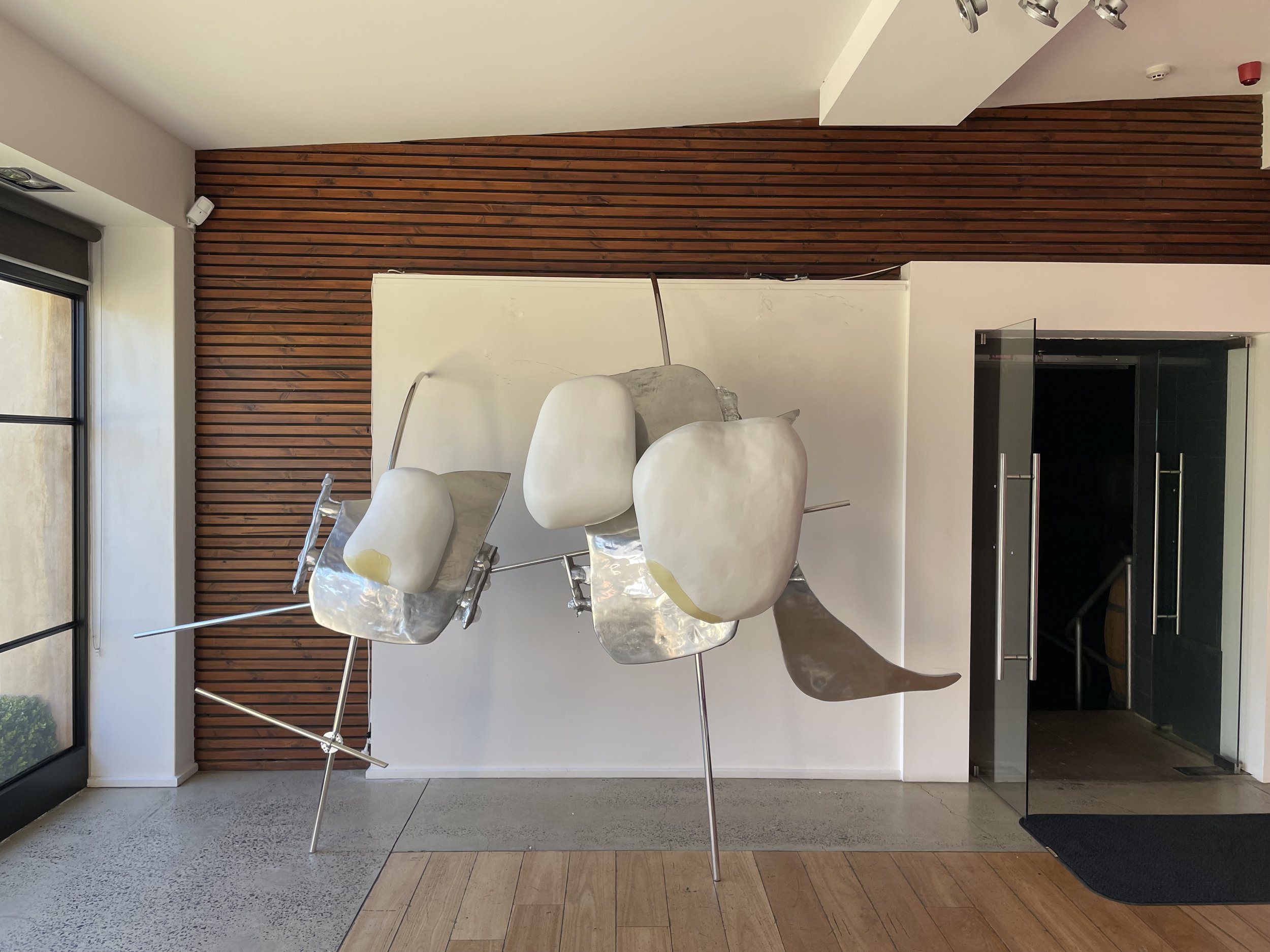 Modern art sculpture of abstract white and metallic forms in a hallway with wood paneling, glass door, and a window.
