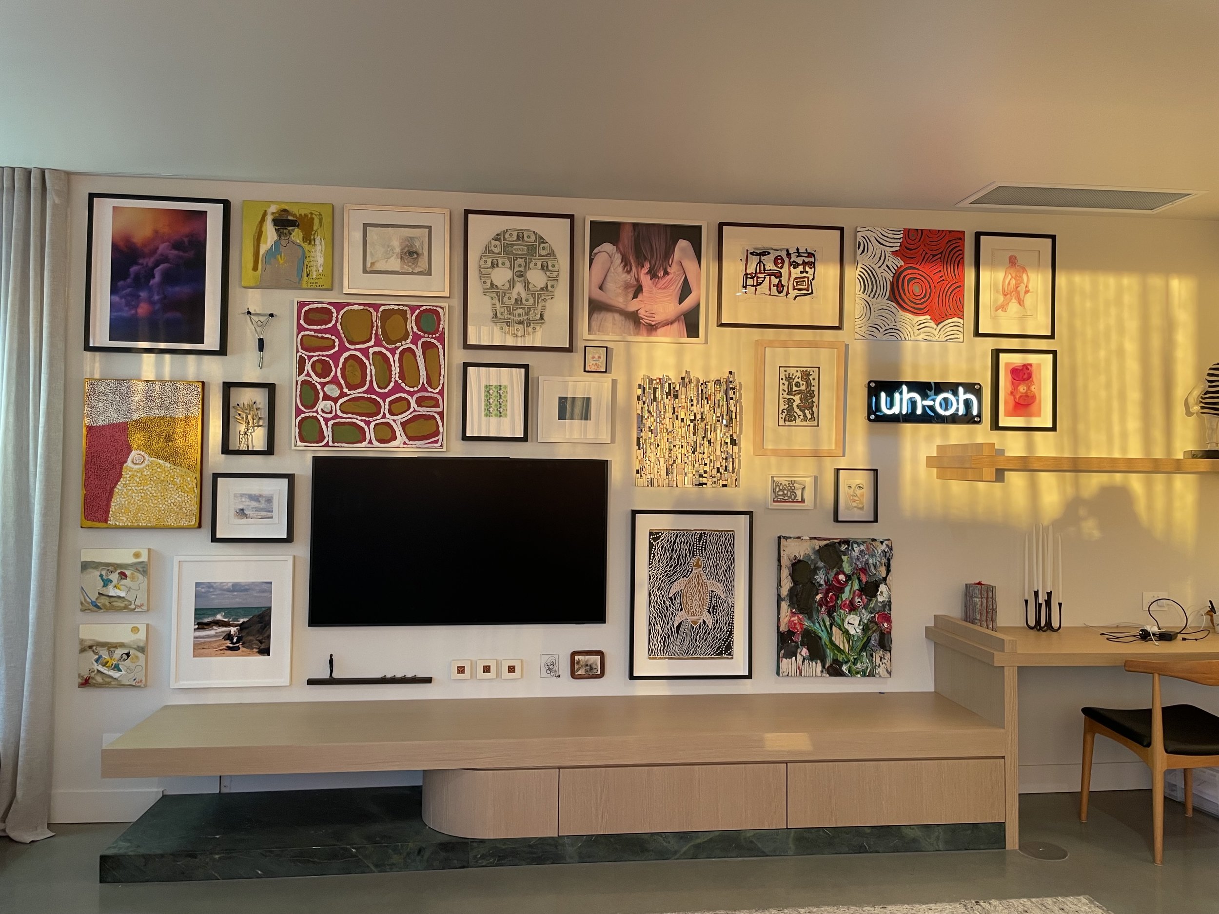A modern living room wall decorated with various framed artworks and a large flat screen TV in the center. To the right, a yellow glow casts shadows on the wall. A neon sign with the text 'uh-oh' is illuminated, and a wooden sideboard holds candles and decorative items.