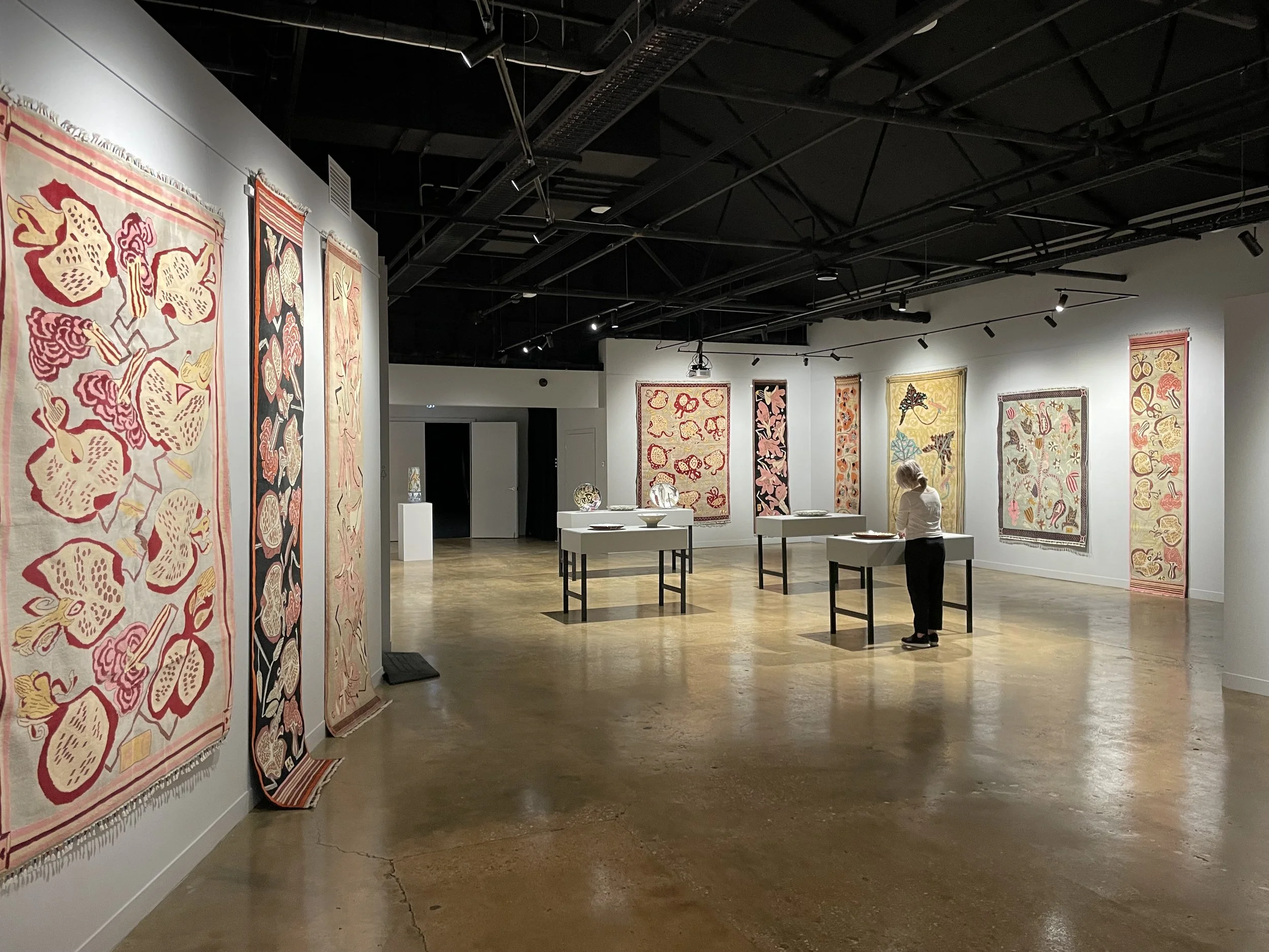 Art gallery with colorful woven tapestries hanging on white walls, several display tables with ceramic pieces, and a visitor observing the artwork. art gallery install, art installation, gallery hang, install of artwork, picture hanging.