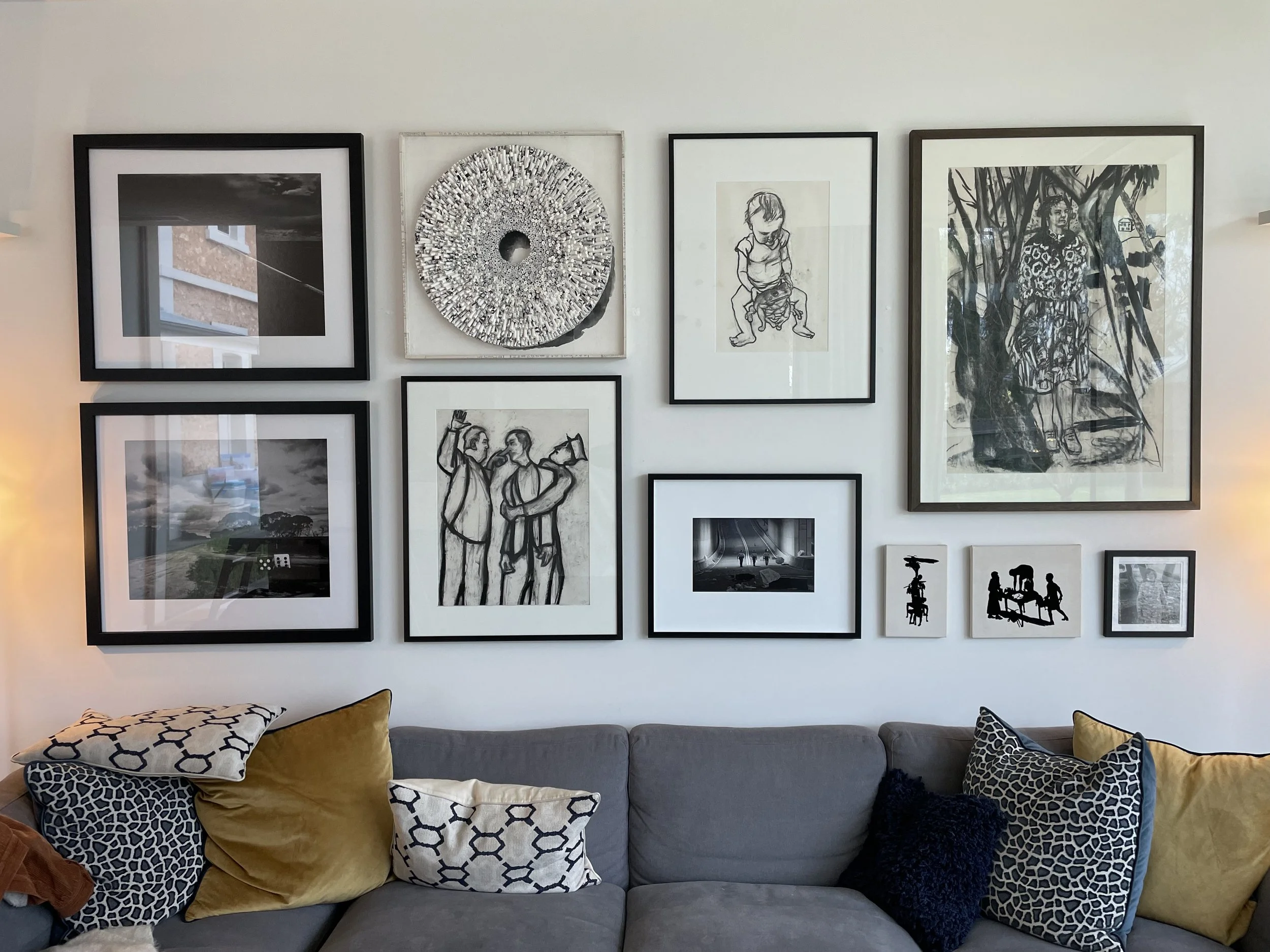 Living room wall decorated with a gallery of black and white framed artwork, with a gray sofa below and patterned pillows in black, white, and yellow.