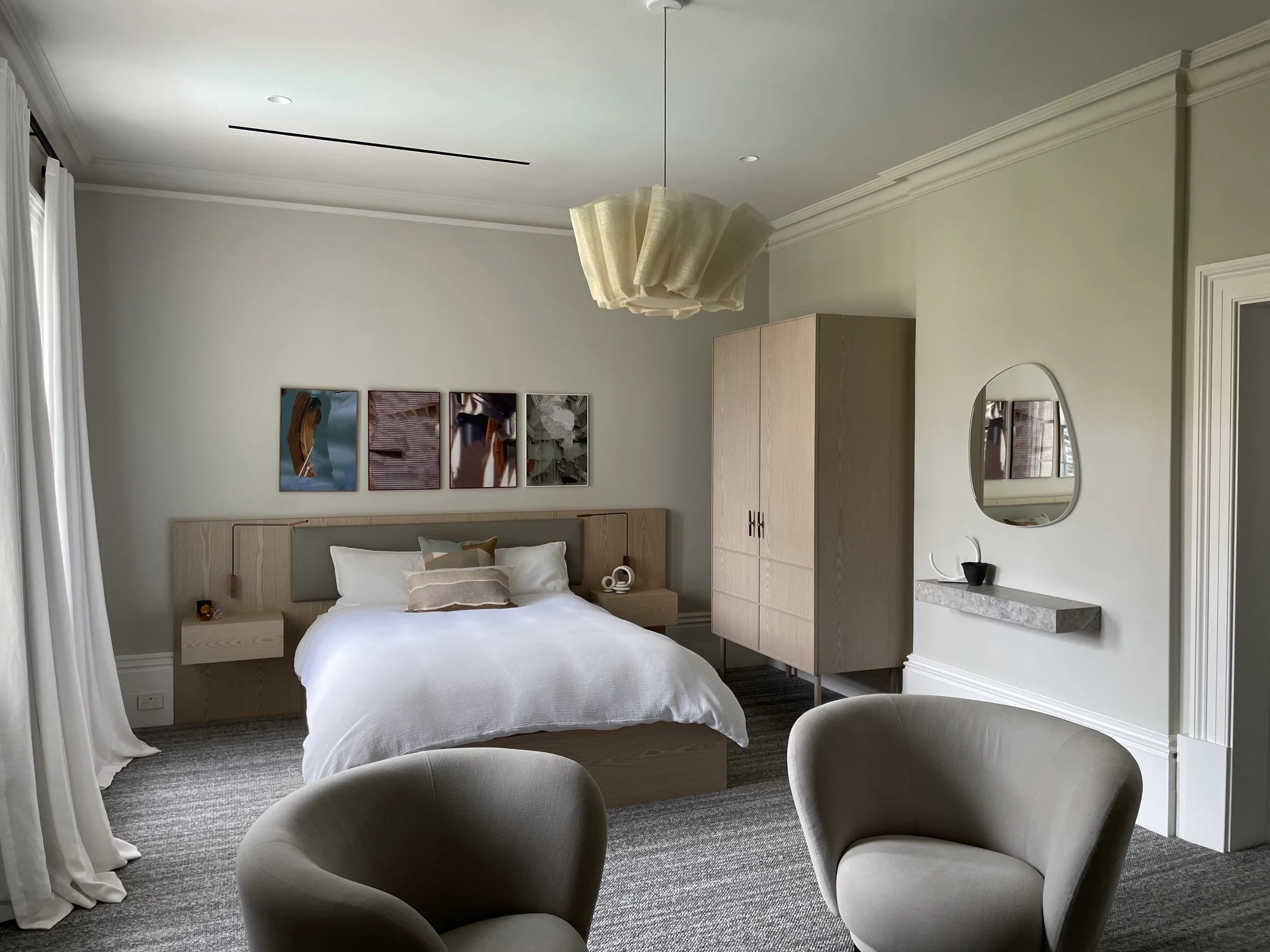 Modern bedroom with a white bed, beige armchairs, a wardrobe, a floating shelf, a mirror, and abstract artwork on the wall.