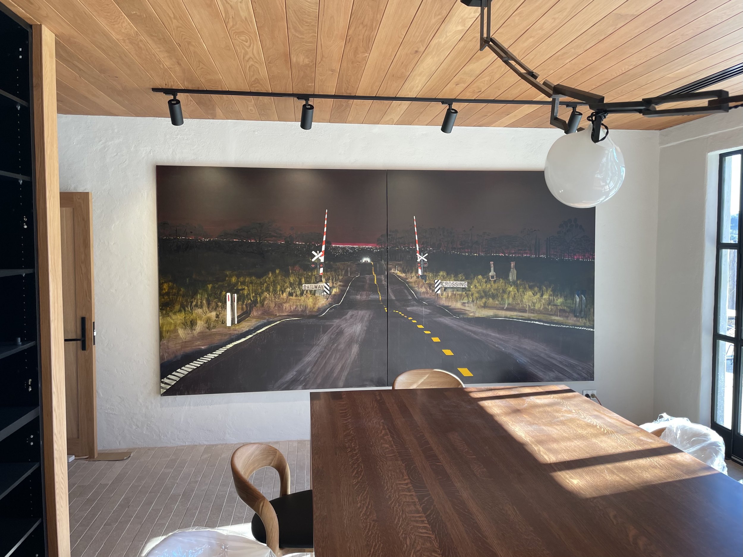 Interior of a room with a large wall art piece of a night-time rural road scene with railroad crossing signs, a wooden table, wooden chairs, track lighting, a pendant light, a window, and a wooden ceiling.