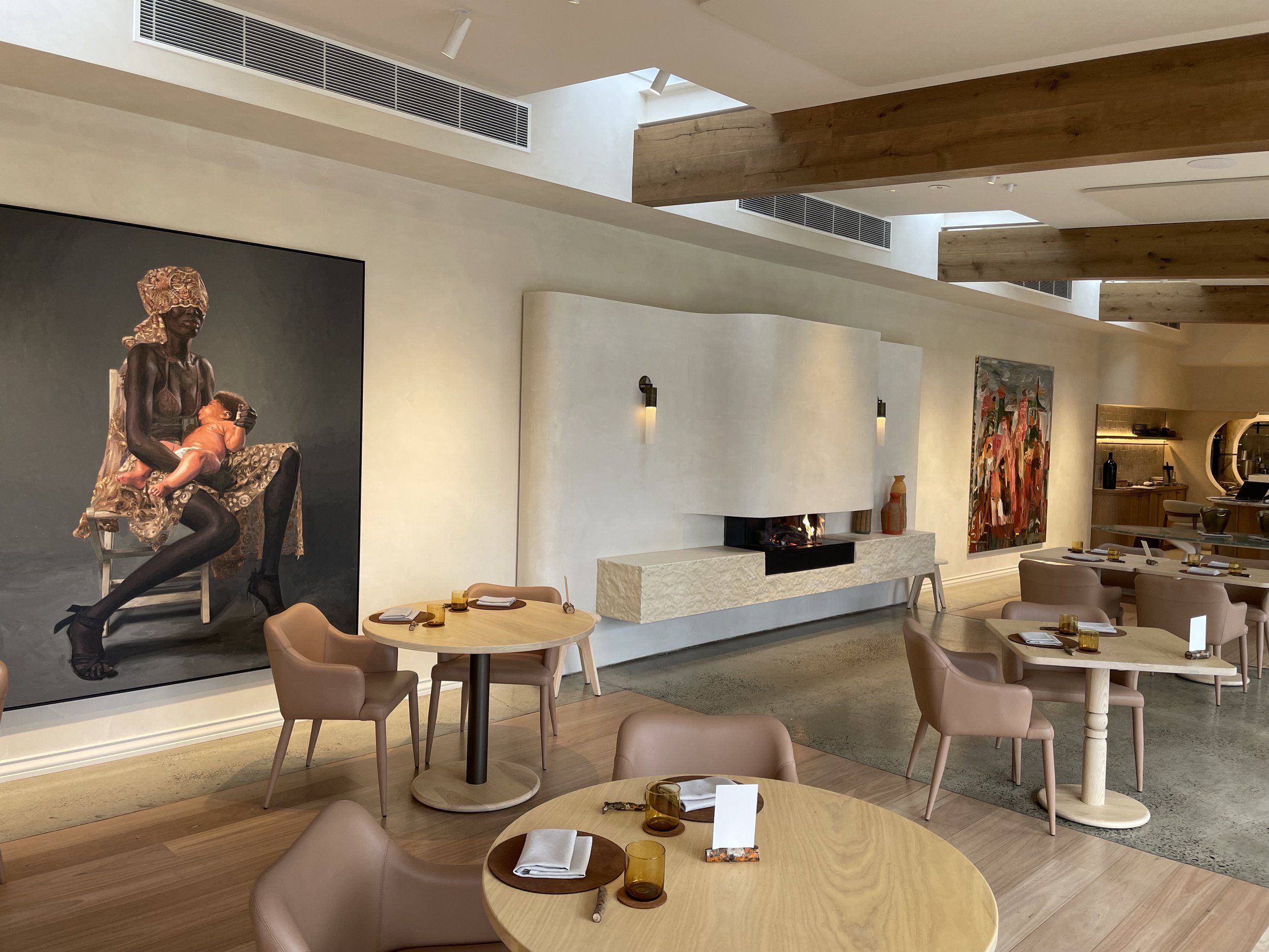 Modern restaurant interior with round wooden tables and pink chairs, wall art including a large painting of a woman holding a child, and a white fireplace on the back wall.