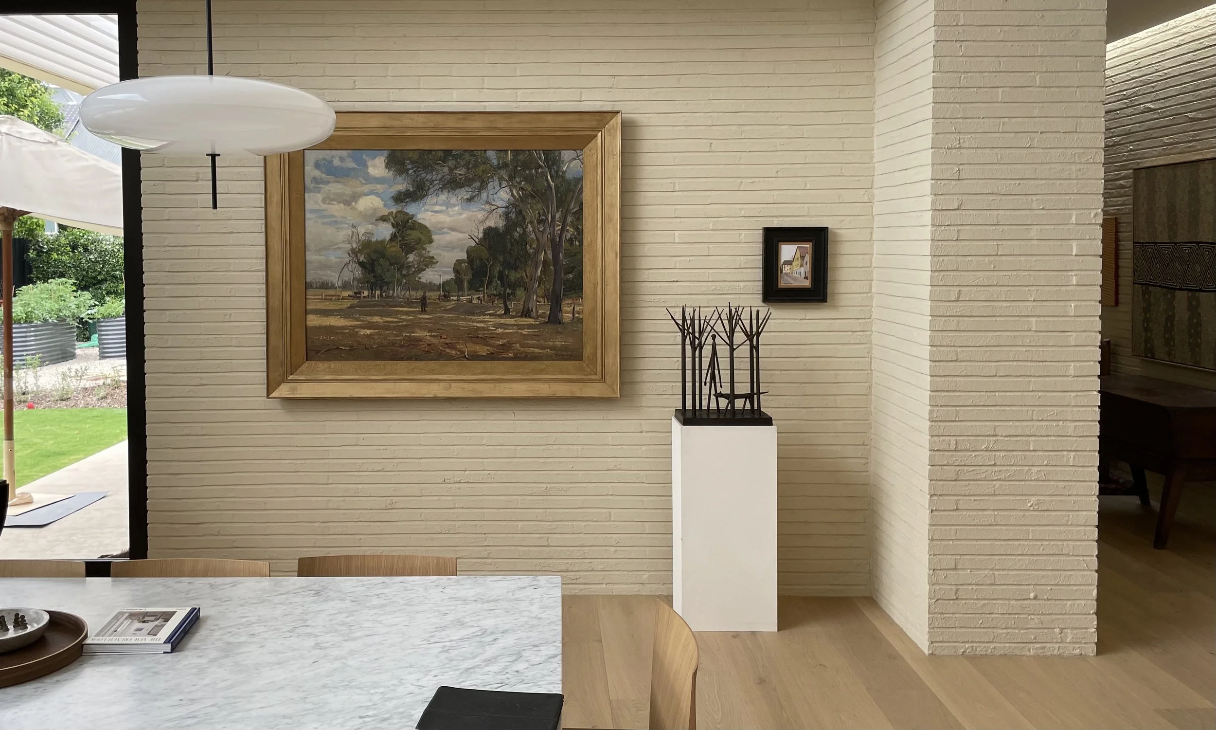 Interior view of a dining area with a marble table, painting of a landscape with trees and sky on the wall, sculpture on a white pedestal, small framed picture, and a modern white hanging light fixture.