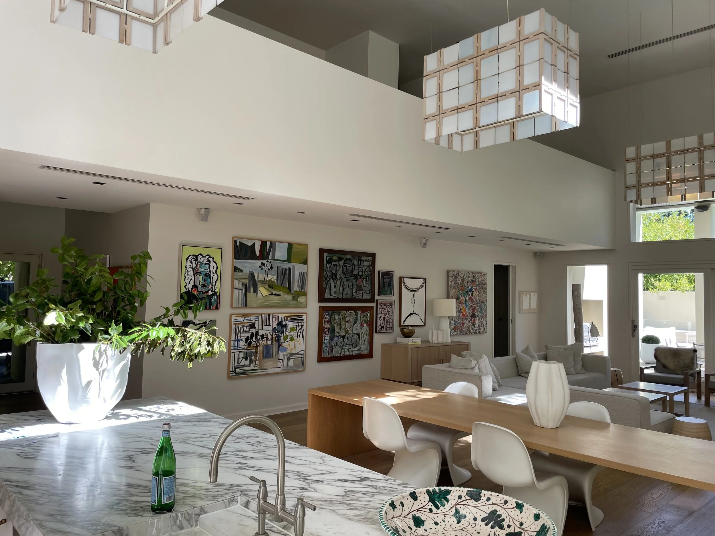 Open-concept living room and kitchen with modern art on the wall, white sofas, large windows, and decorative lighting fixtures.