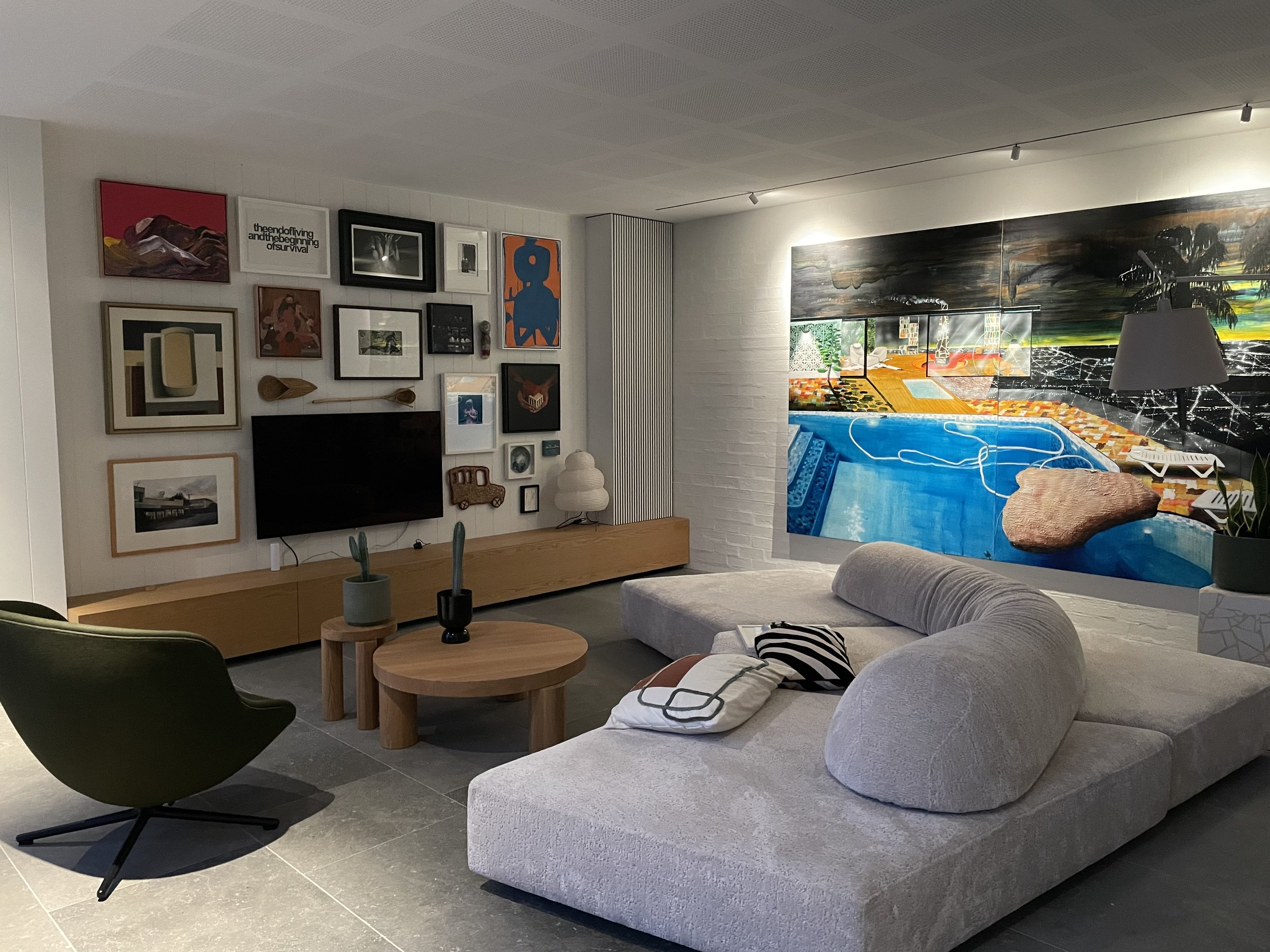 Modern living room with gallery wall, large abstract painting, curved beige sofa with black and white pillows, round wooden coffee table with small potted plants, dark green armchair, wall mounted TV, small cabinet, and a large mural of a pool scene.