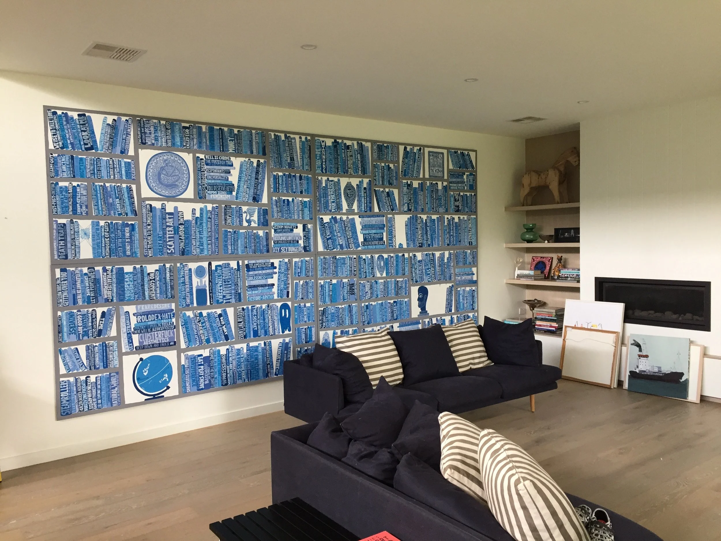 Living room with modern decor, featuring a large blue and white art piece made of books on the wall, a black sofa with striped and black cushions, wooden flooring. art installation, art install, picture hanging. interior design.