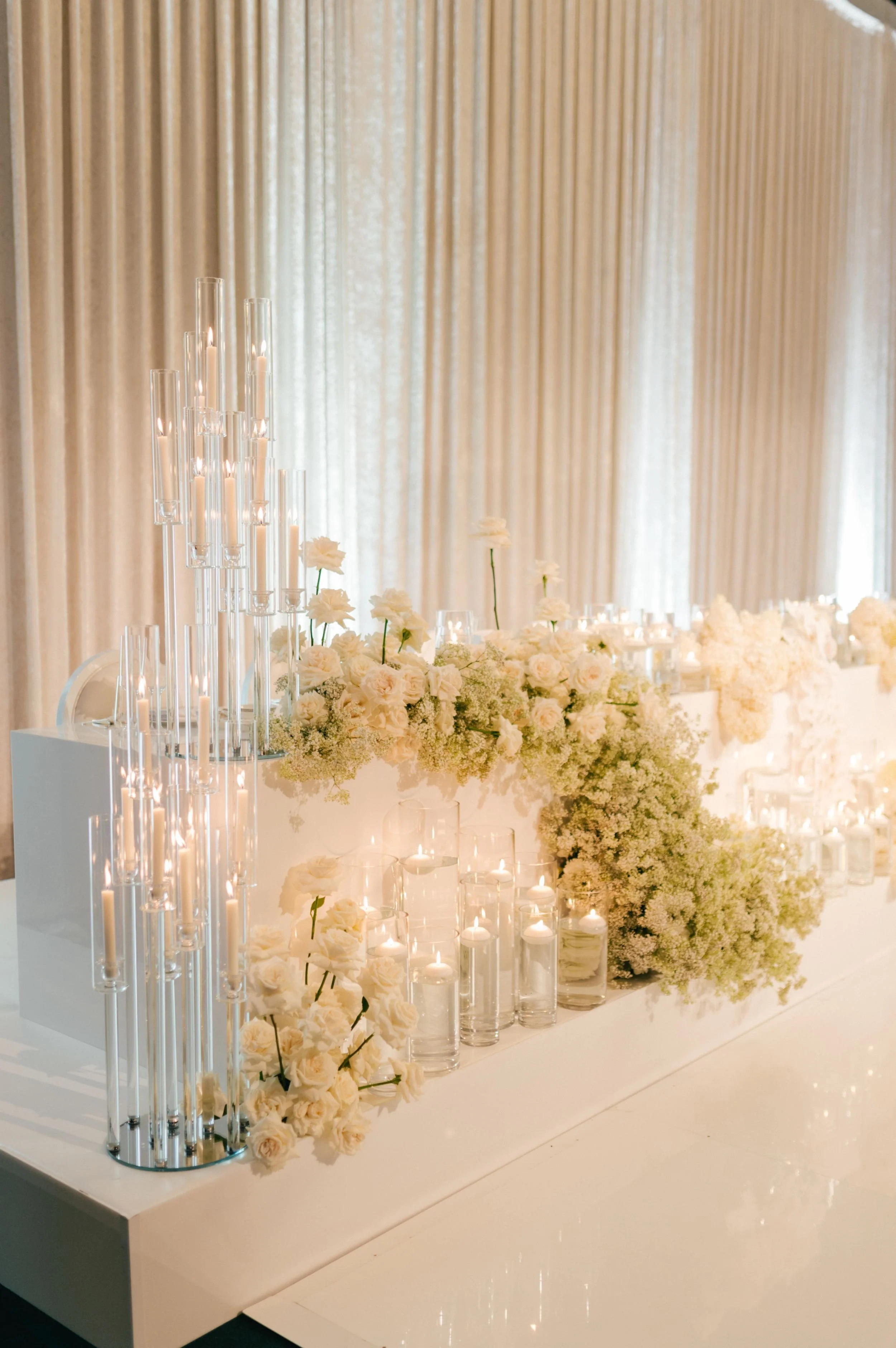 Photo by Kenneth Lim Photography.  Elegant display of white candles in glass holders and a large arrangement of white roses and other flowers with a beige curtain backdrop.  Candle styling by Candlescapes, Perth Candle Styling & Hire.