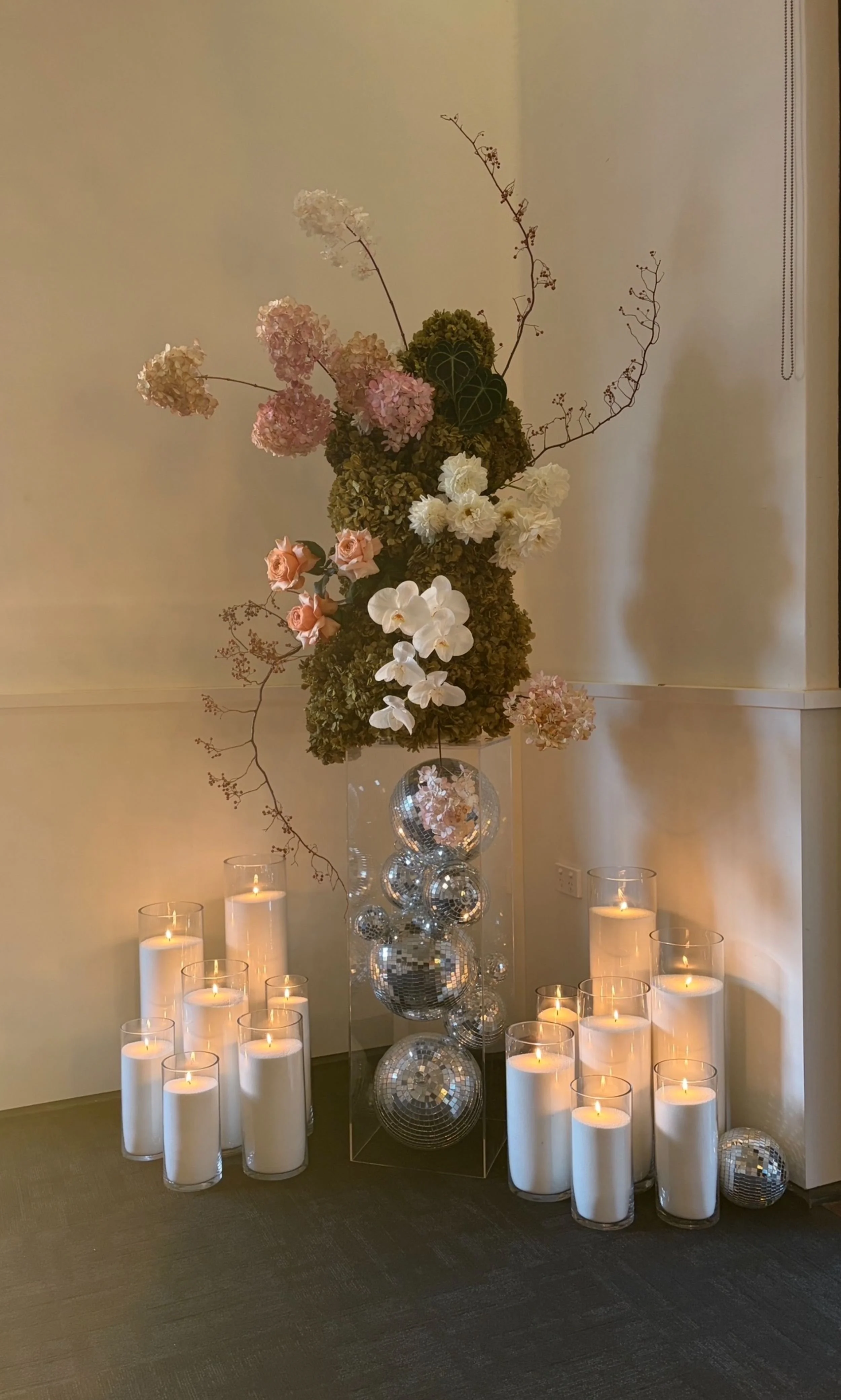 A floral arrangement with pink, white, and green flowers and branches on a transparent stand, surrounded by lit white candles and disco ball decorations.