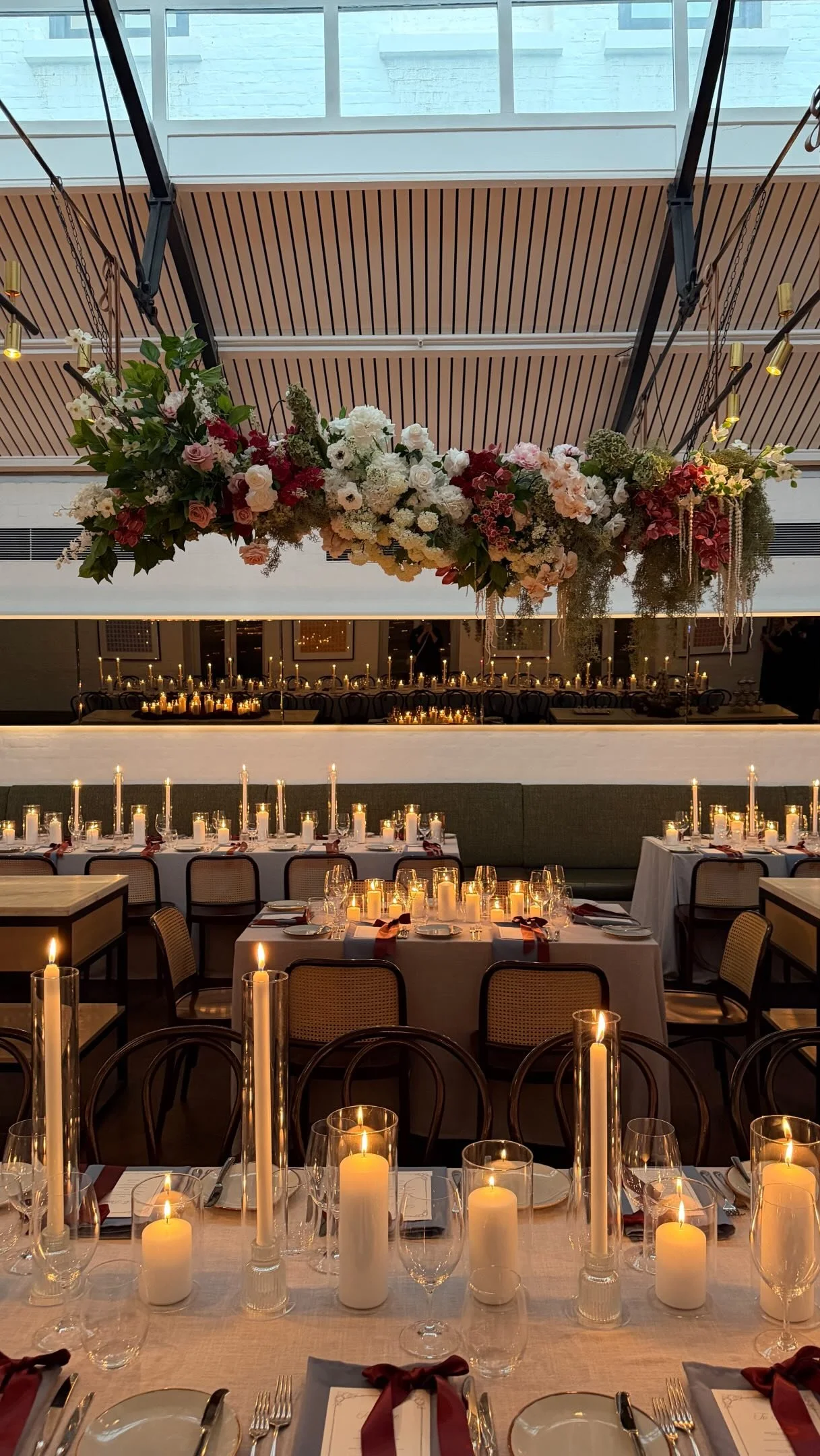 Elegant dining setup with long tables covered with white tablecloths, surrounded by chairs, decorated with numerous candles of varying heights in glass holders, and set with plates, glassware, and cutlery. A large floral arrangement of white, pink, and red flowers with green foliage hangs from the ceiling above the tables.