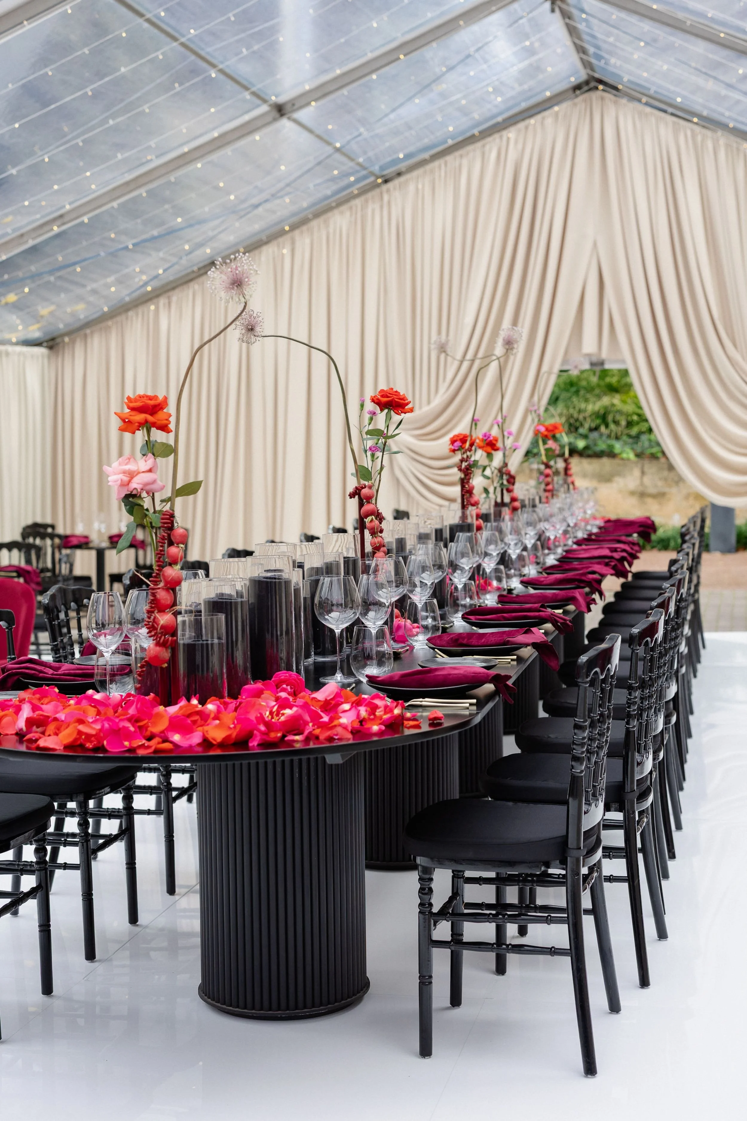 Photo by Tessa Kit Photography.  A long banquet table decorated with pink, red, and orange flowers and rose petals, set with wine glasses, black plates, and burgundy napkins, under a white tent with draped curtains and string lights.