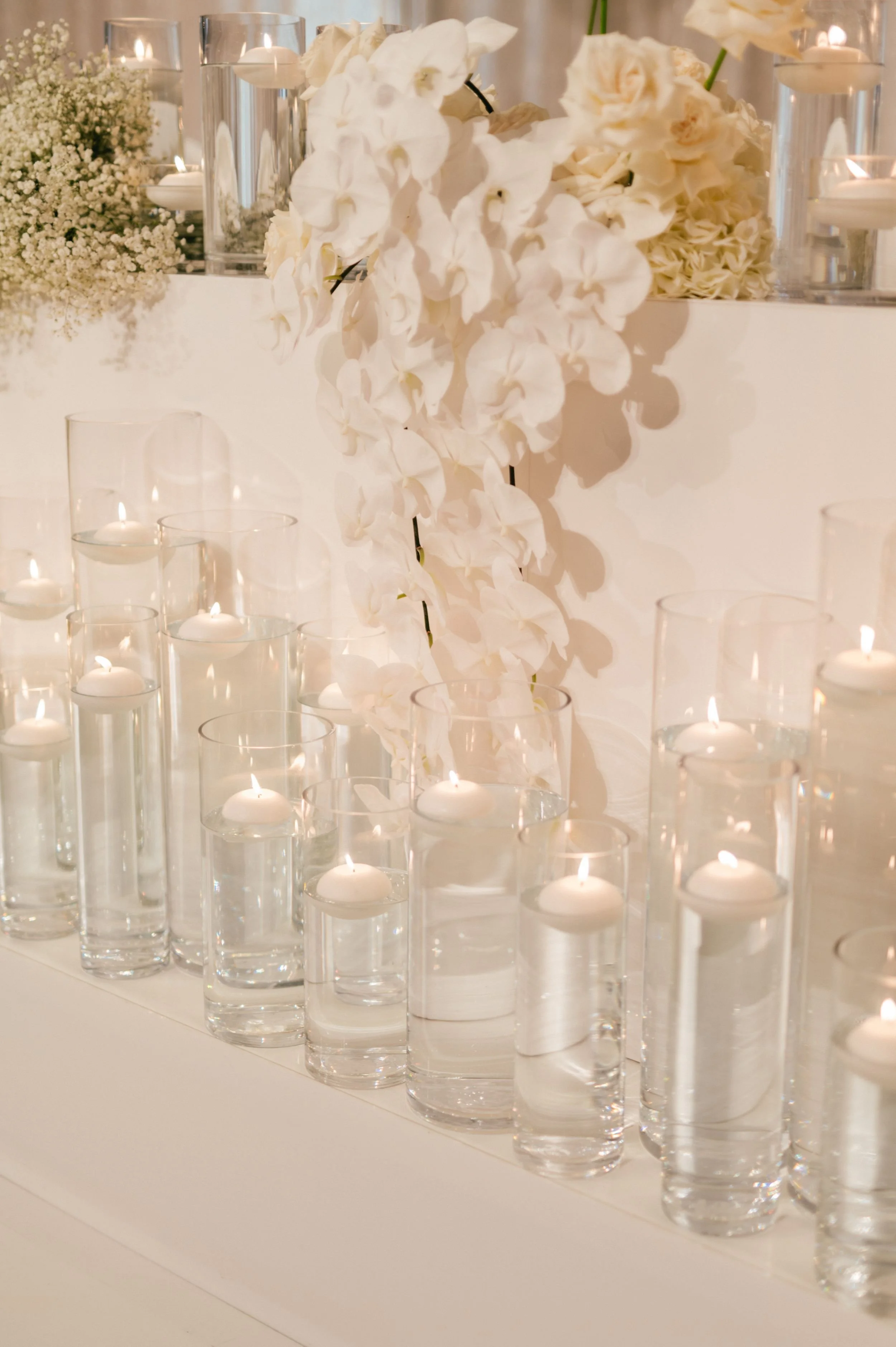 Photo by Kenneth Lim Photography.  White candles in glass holders arranged in rows, with white flowers and candlelit vases in the background, creating an elegant decorative display.
