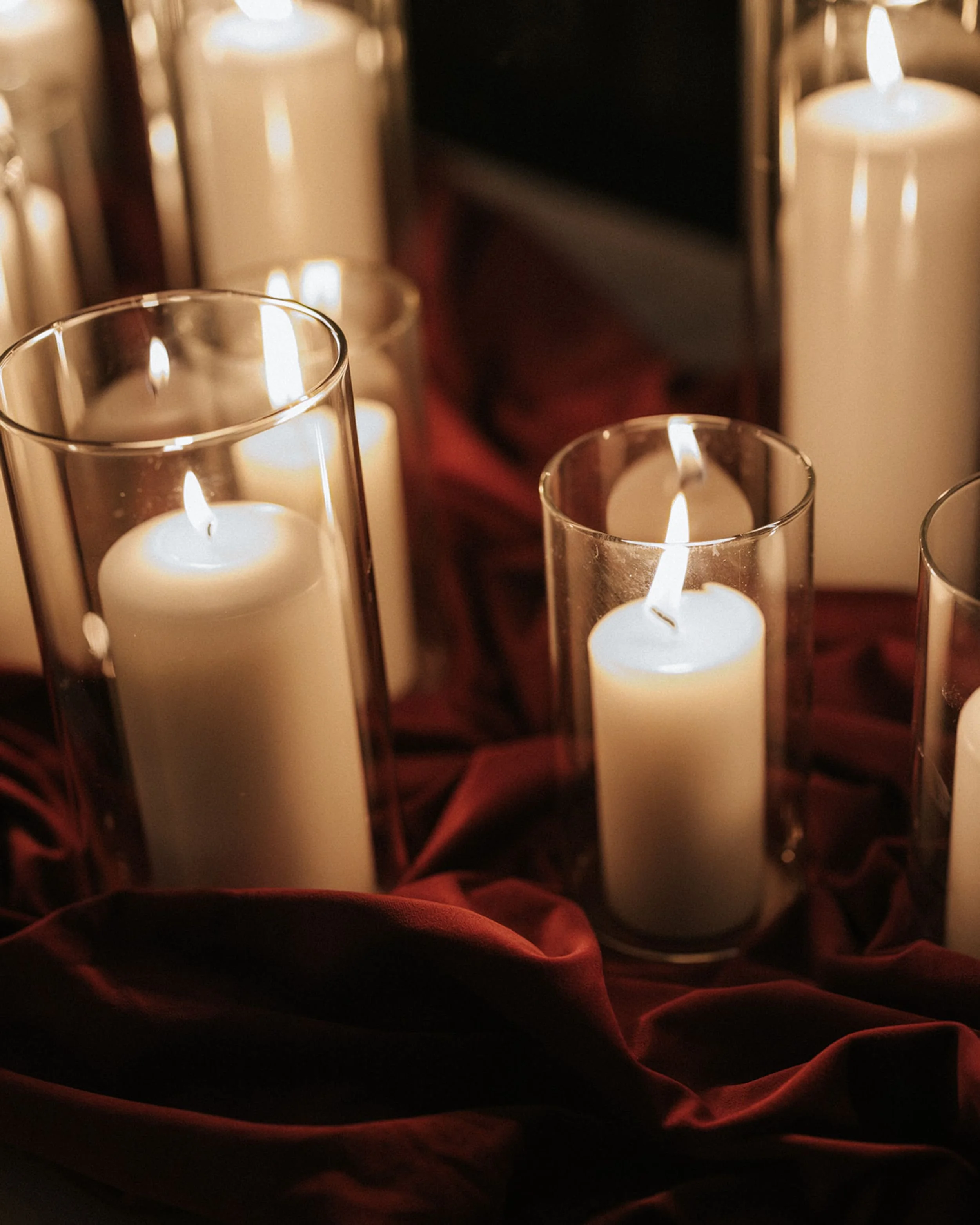 Photo by Sunny Bishop Photography.  Multiple lit white candles in glass holders, placed on a surface with a red cloth underneath, creating a warm, cosy ambiance.  Candle styling by Candlescapes, Perth Candle Styling & Hire.