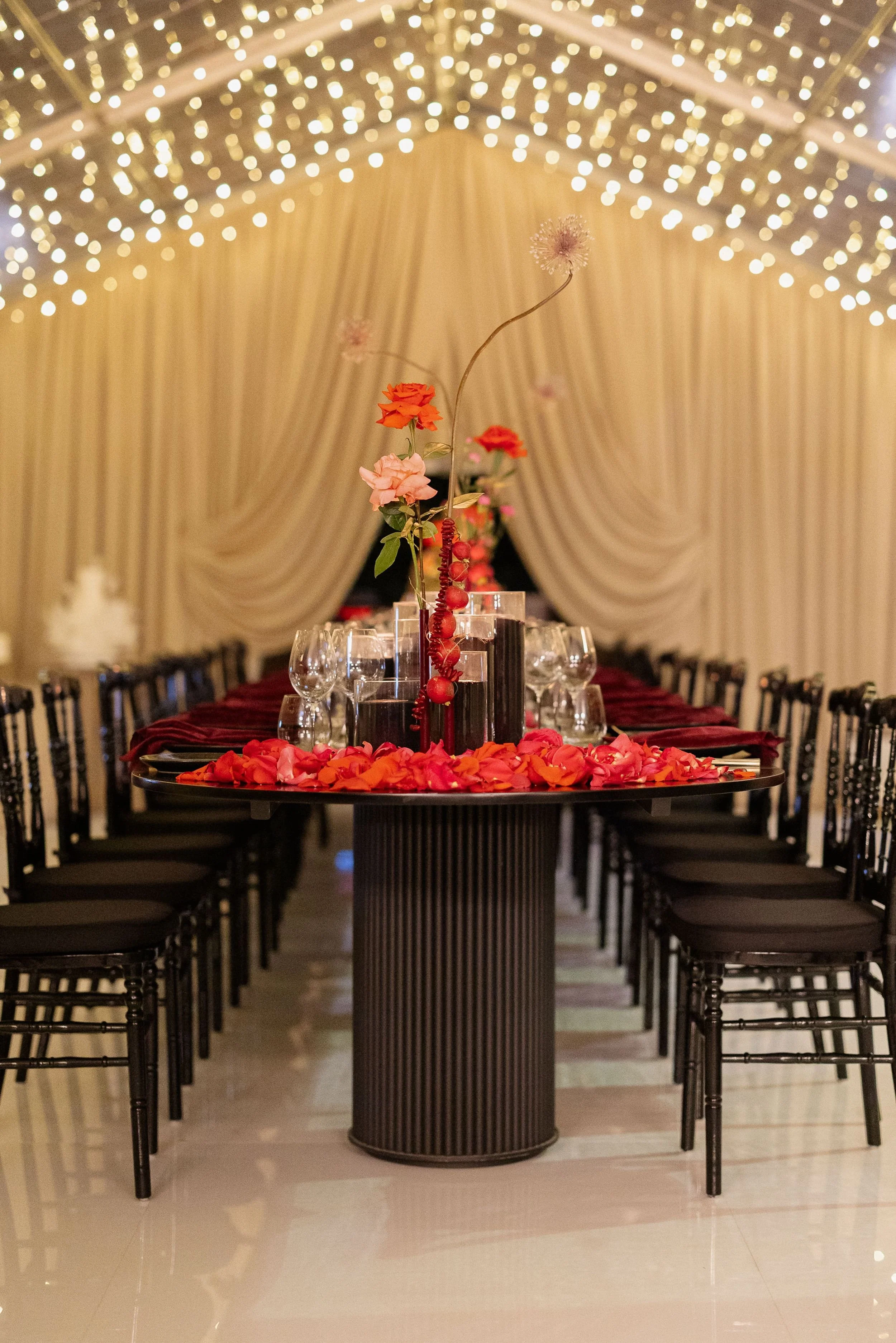 Photo by Tessa Kit Photography.   Elegantly decorated banquet table with a floral centrepiece consisting of pink, orange, and purple flowers, surrounded by scattered rose petals, under a canopy of string lights with beige drapery in the background.  