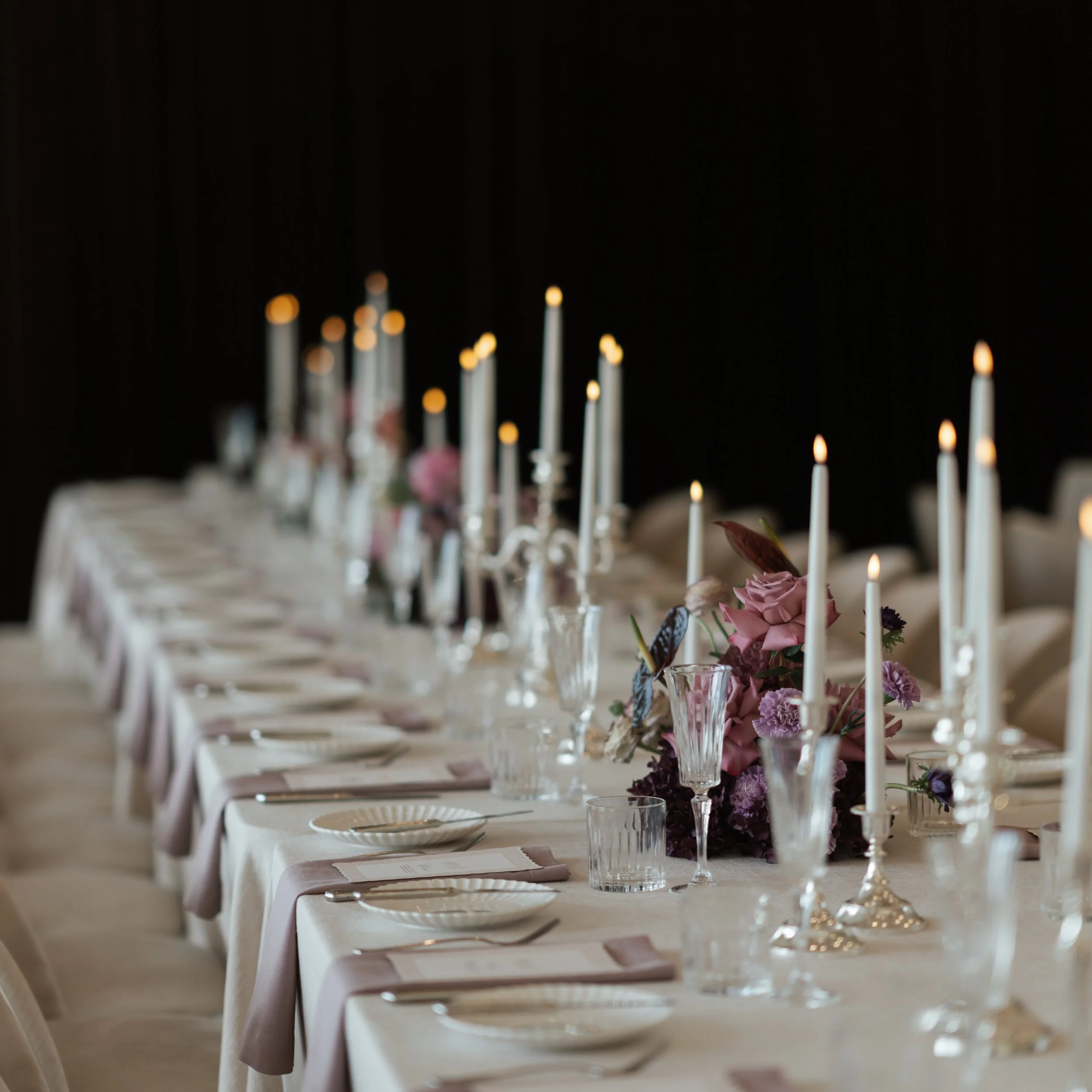 A long dining table set for a celebration or event, decorated with pink flowers, tall white candles, and elegant glassware, with chairs on either side and a dark background.