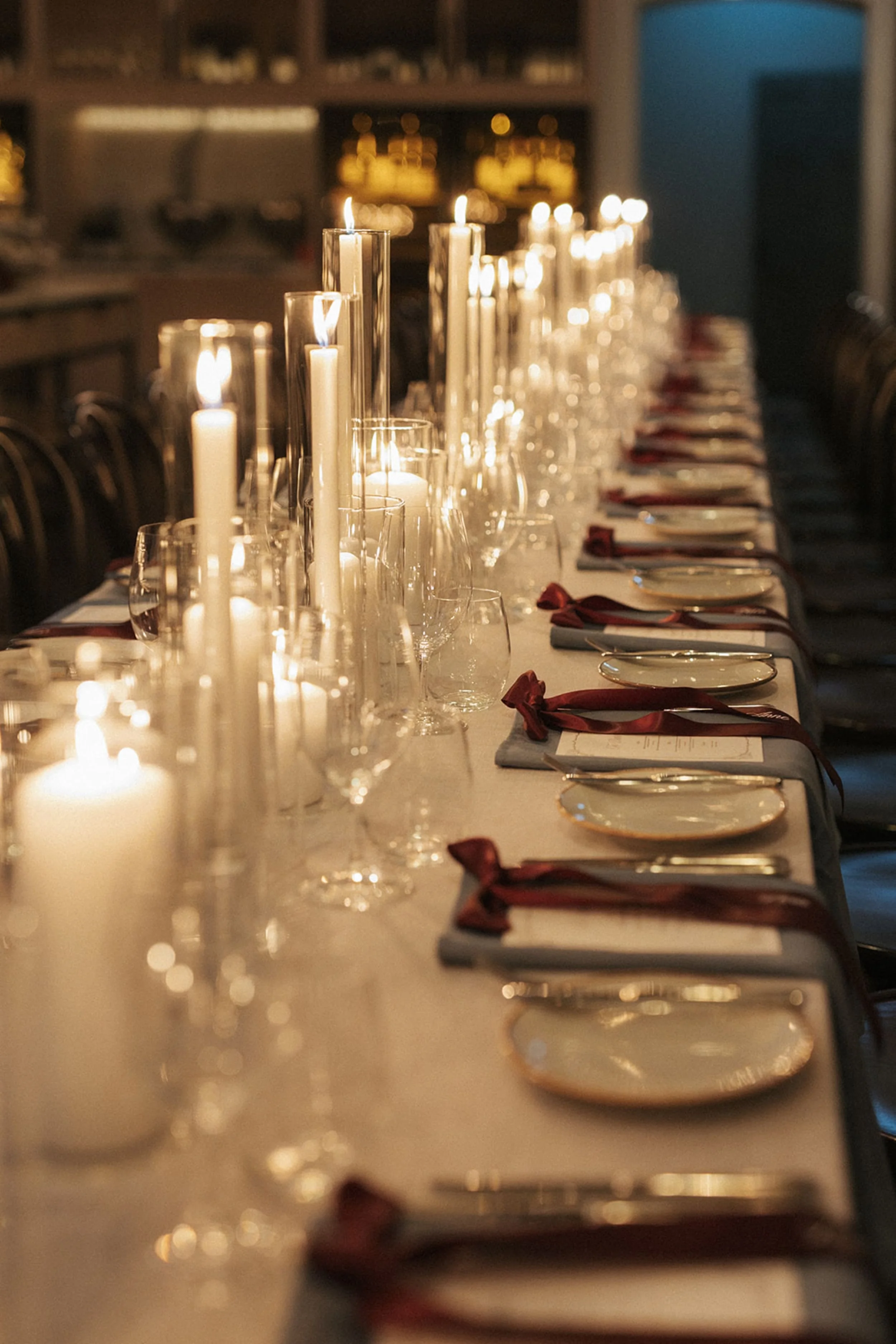 Photo by Sunny Bishop Photography.  A long dining table set for a formal dinner, with lit candles in glass holders, wine glasses, plates, and red cloth napkins on each place setting, in a dimly lit room.