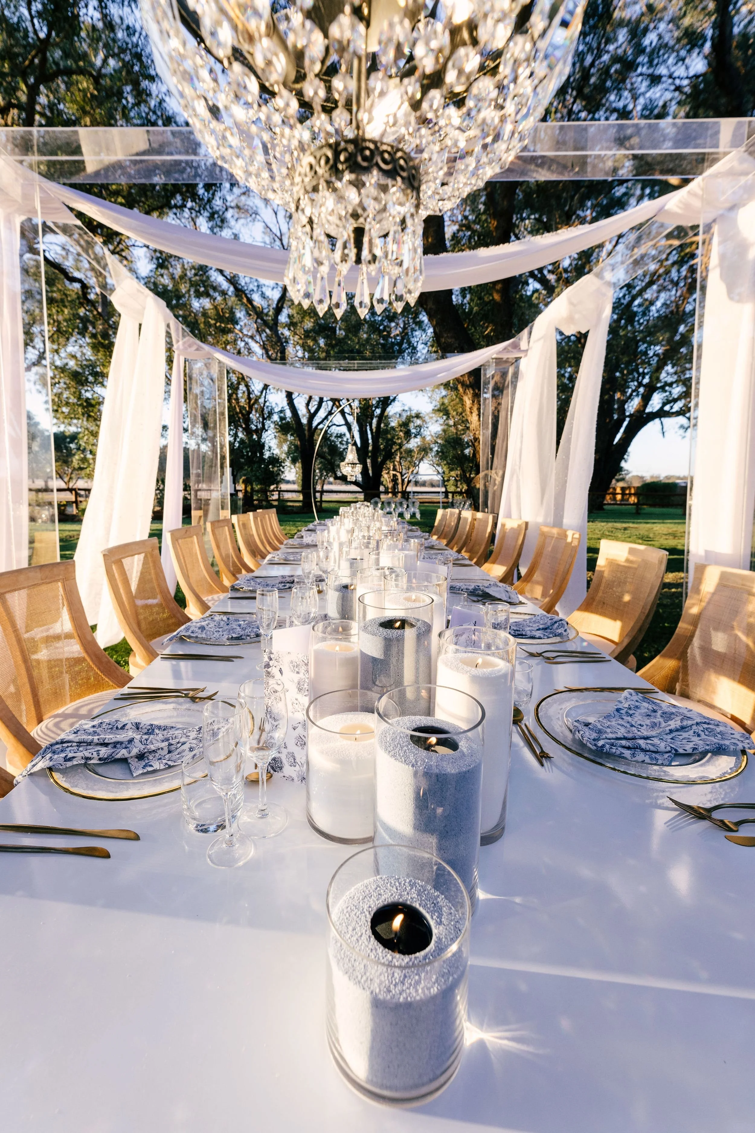 Photo by Compose Photography.  Elegant outdoor dining setup with a long white table, crystal chandeliers hanging overhead, and candles in glass holders along the center of the table. White draped fabric and trees are visible in the background.