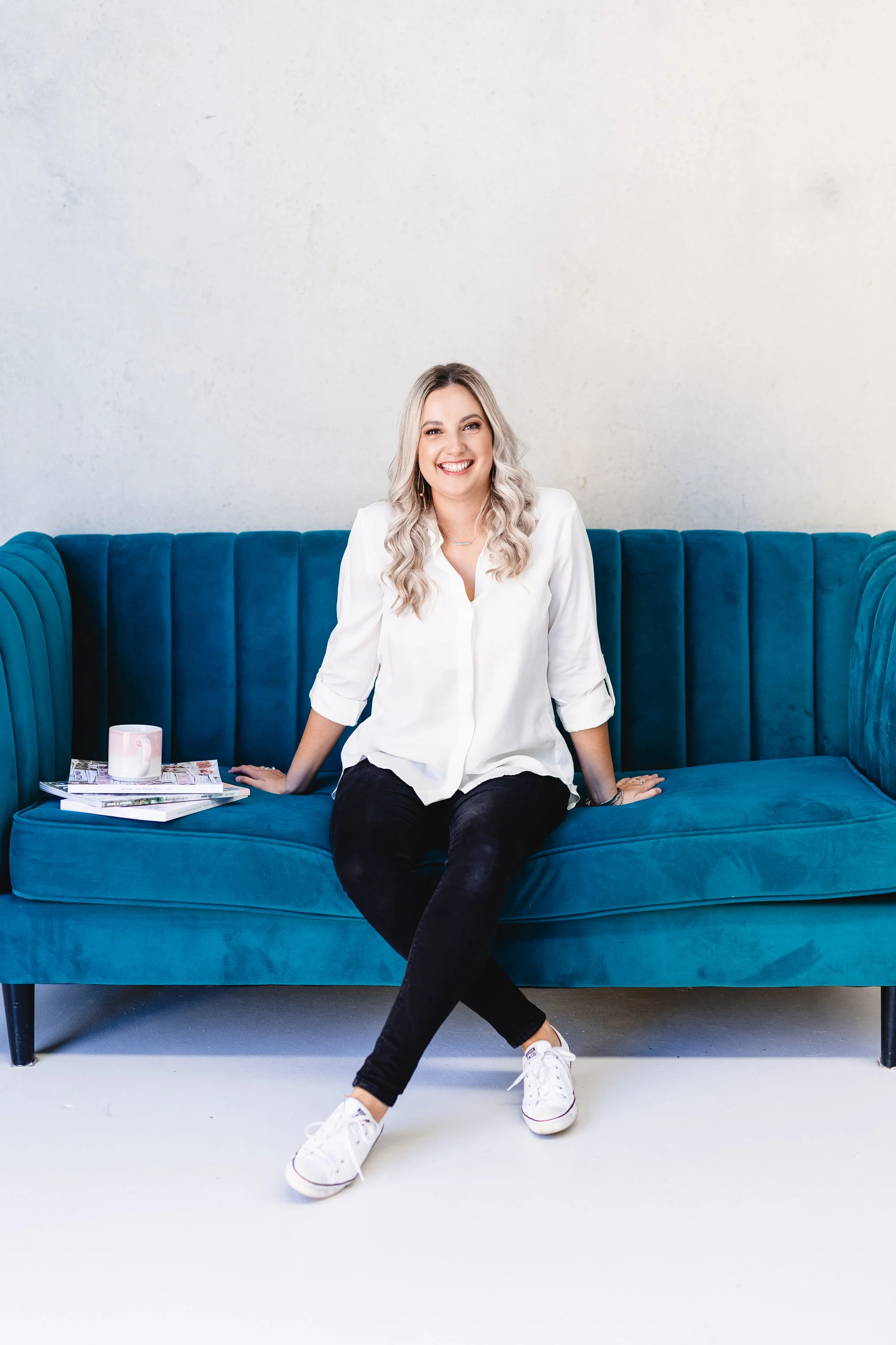 Aimee Shadbolt, of Candlescapes, Perth.  She has blonde wavy hair, wearing a white shirt, black pants, and white sneakers, and is sitting on a blue velvet couch against a plain white wall. There are magazines and a mug on the left side of the couch.