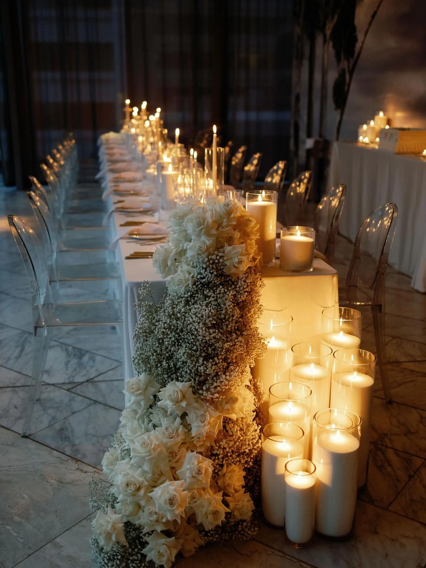 Elegant banquet table decorated with white roses and baby's breath floral arrangement, surrounded by tall white candles in glass holders, set in a dimly lit room.  Candle styling by Candlescapes, Perth Candle Styling & Hire.