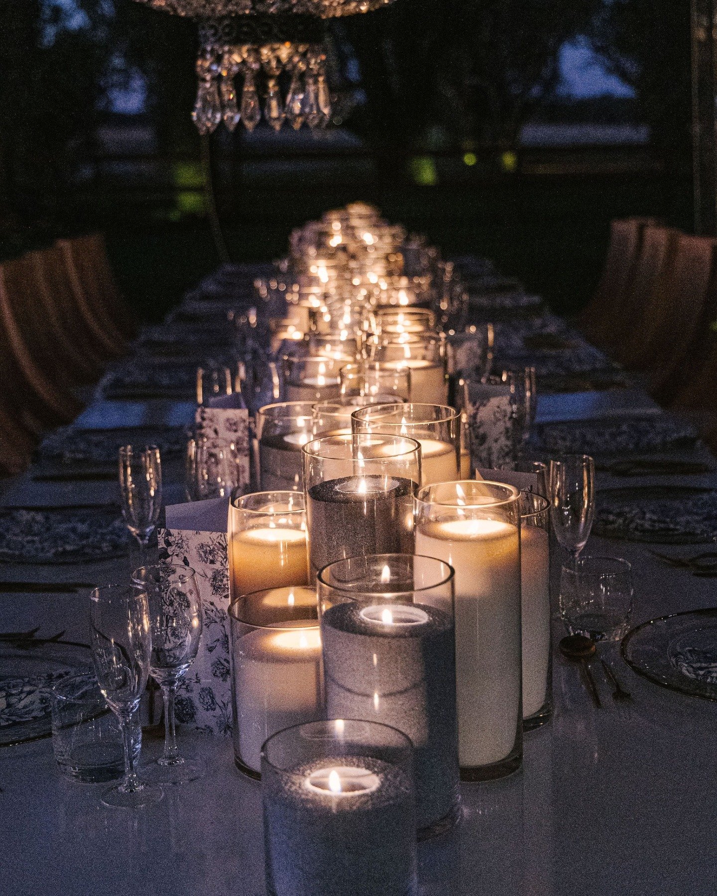 A table decorated with numerous lit candles of varying heights, surrounded by empty glasses and set for a meal, with a dark background. Candle styling by Candlescapes, Perth Candle Styling & Hire.