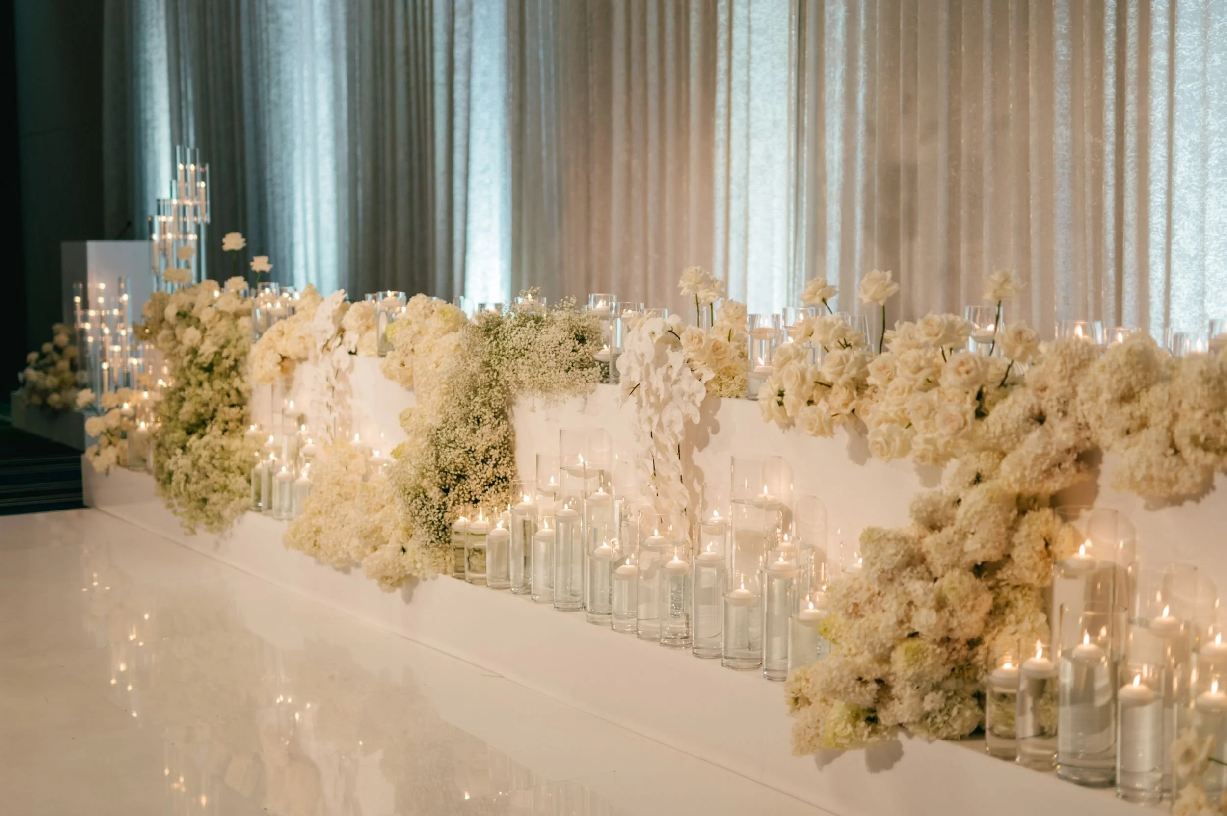 Photo by Kenneth Lim Photography.  Elegant wedding or event table decor with white flowers, roses, and baby's breath, illuminated by candles in tall glass holders, set against a backdrop of beige curtains.