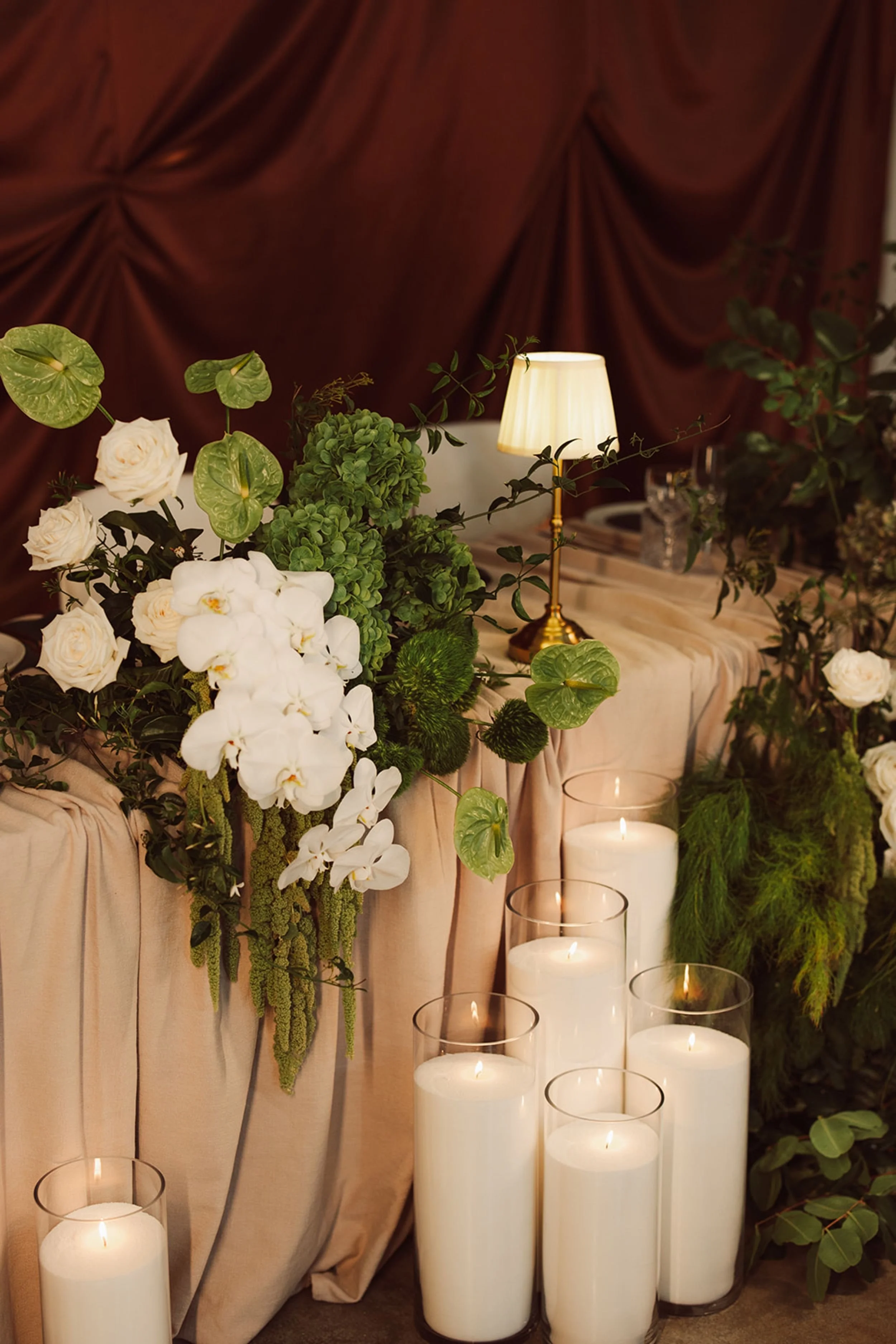 Photo by Jamie Lee Photography.   A floral decoration with white roses, green hydrangeas, orchids, and other greenery, arranged on a table with candles and draped cloth in the background, for an event or celebration.