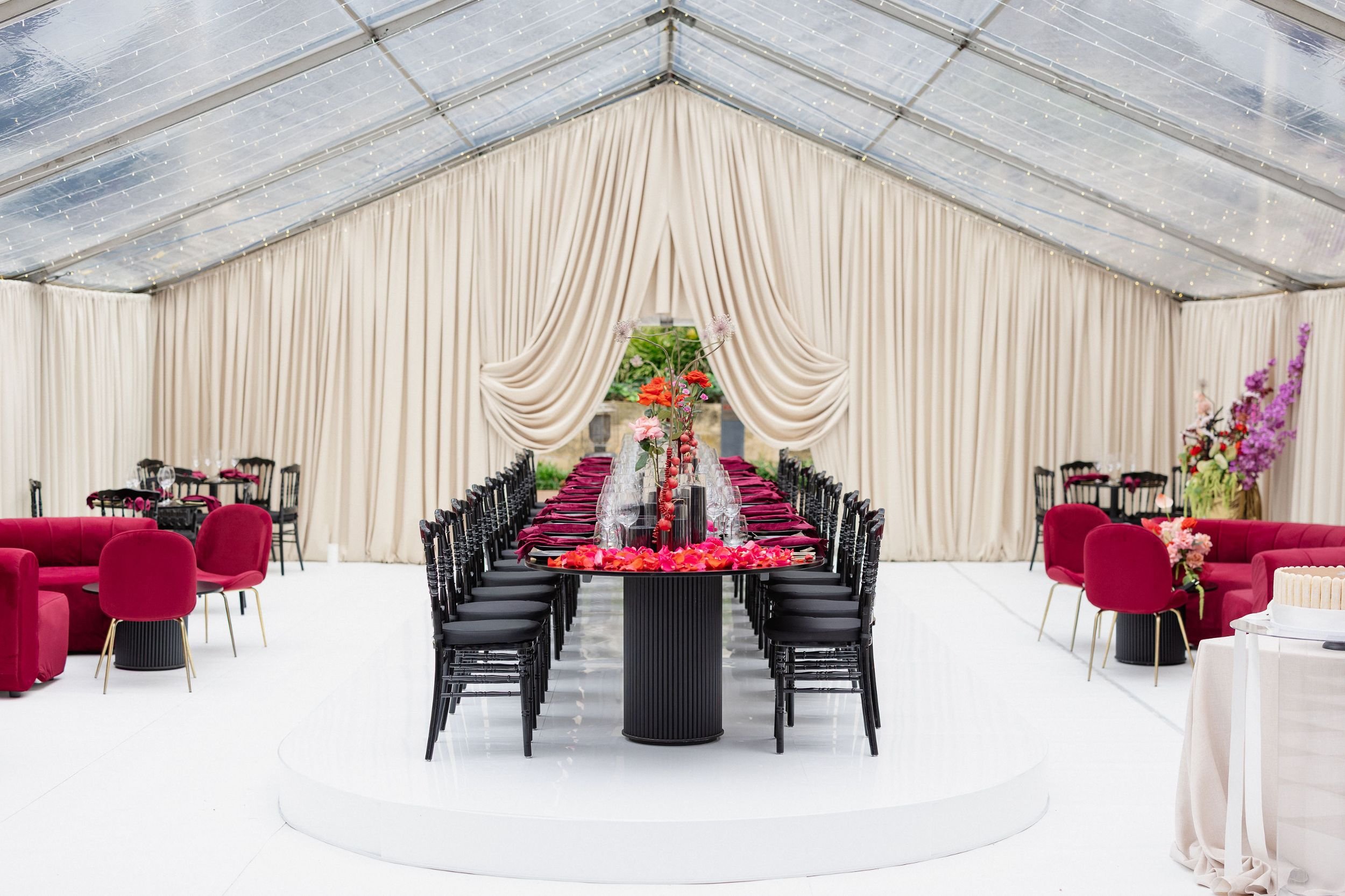 Photo by Tessa Kit Photography. Elegant event tent with long dining table decorated with flowers, surrounded by black chairs, cream draped curtains, and string lights on the ceiling.