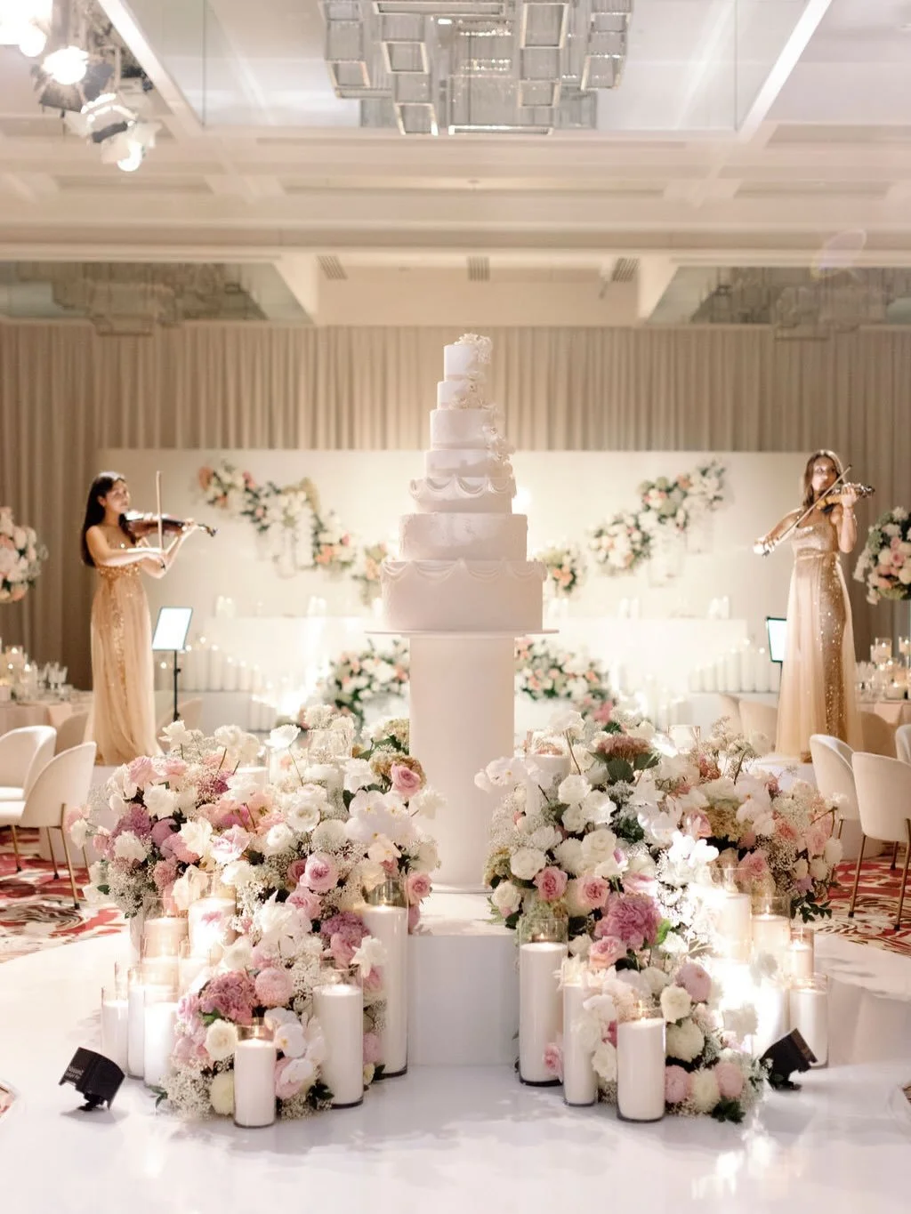 Elegant wedding reception setup with a tall white wedding cake on a floral decorated table, surrounded by pink and white flowers and candles, with two musicians playing violins in the background.  Candle styling by Candlescapes, Perth Candle Styling