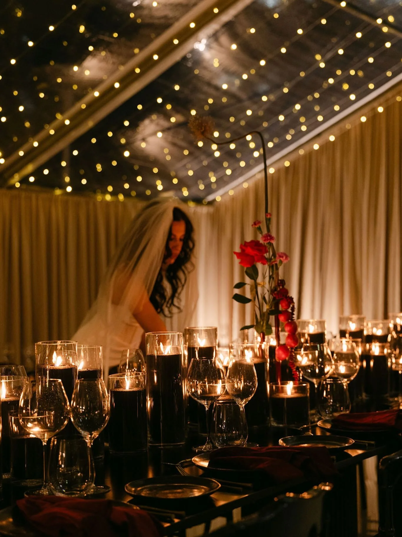 A woman in a wedding dress and veil decorating a table with candles, flowers, and glassware in an elegant setting with a ceiling decorated with string lights.  Candle styling by Candlescapes, Perth Candle Styling & Hire.